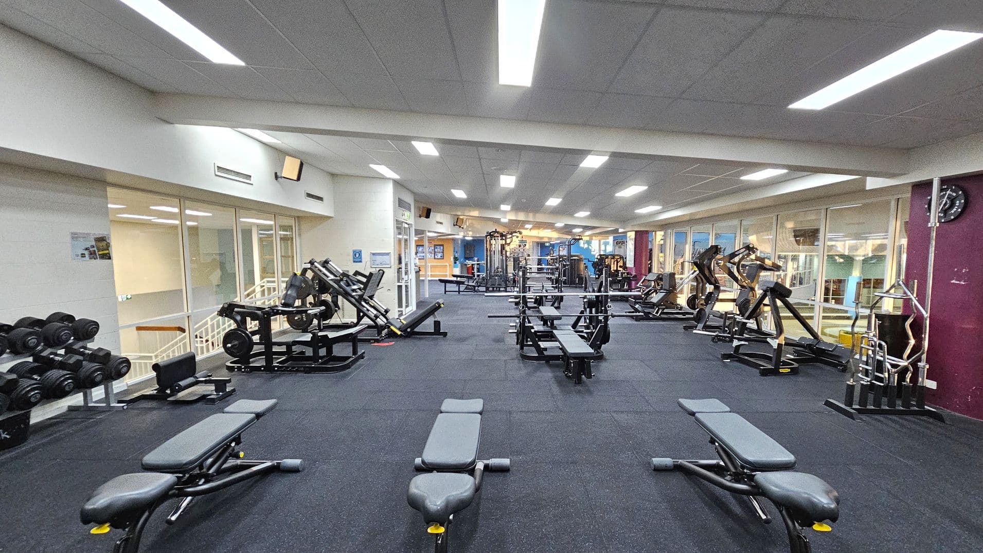 A spacious gym with a dark carpeted floor and a white ceiling with fluorescent lights. In the foreground, there are several weight benches. To the left, a rack of dumbbells is visible. The middle area features various weight machines and benches. On the right, more weight machines are lined up against a wall with large windows. A clock is mounted on the wall to the right. The background shows additional gym equipment and a reception area with blue and orange walls.