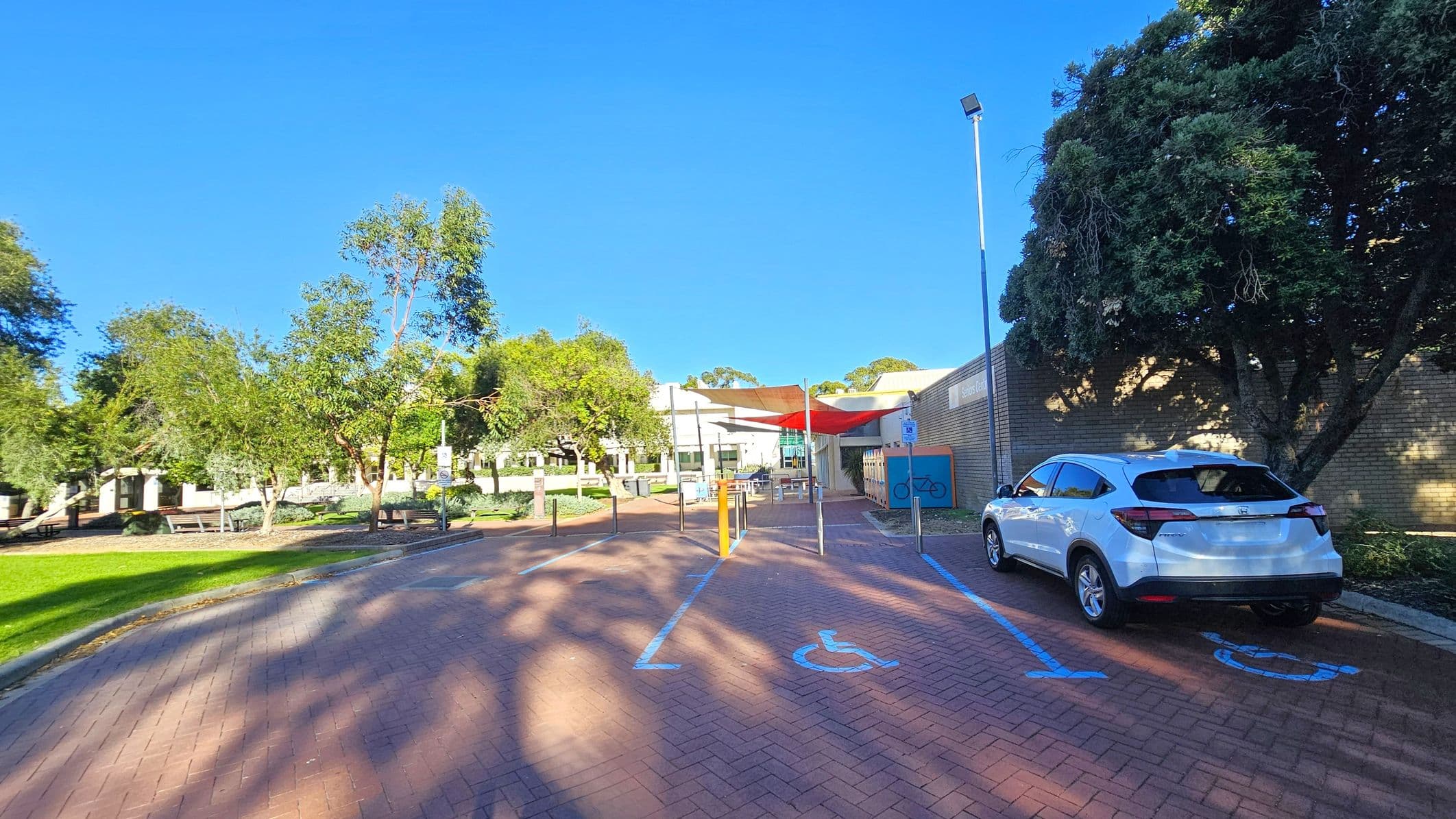 A paved area with designated disabled parking spaces marked in blue. A white car is parked on the right. A brick building with a bicycle sign is on the right, partially shaded by a large tree. In the centre, a red shade structure covers a seating area. On the left, there are green lawns and several trees, with a pathway leading to a white building in the background. A tall streetlamp is near the car.