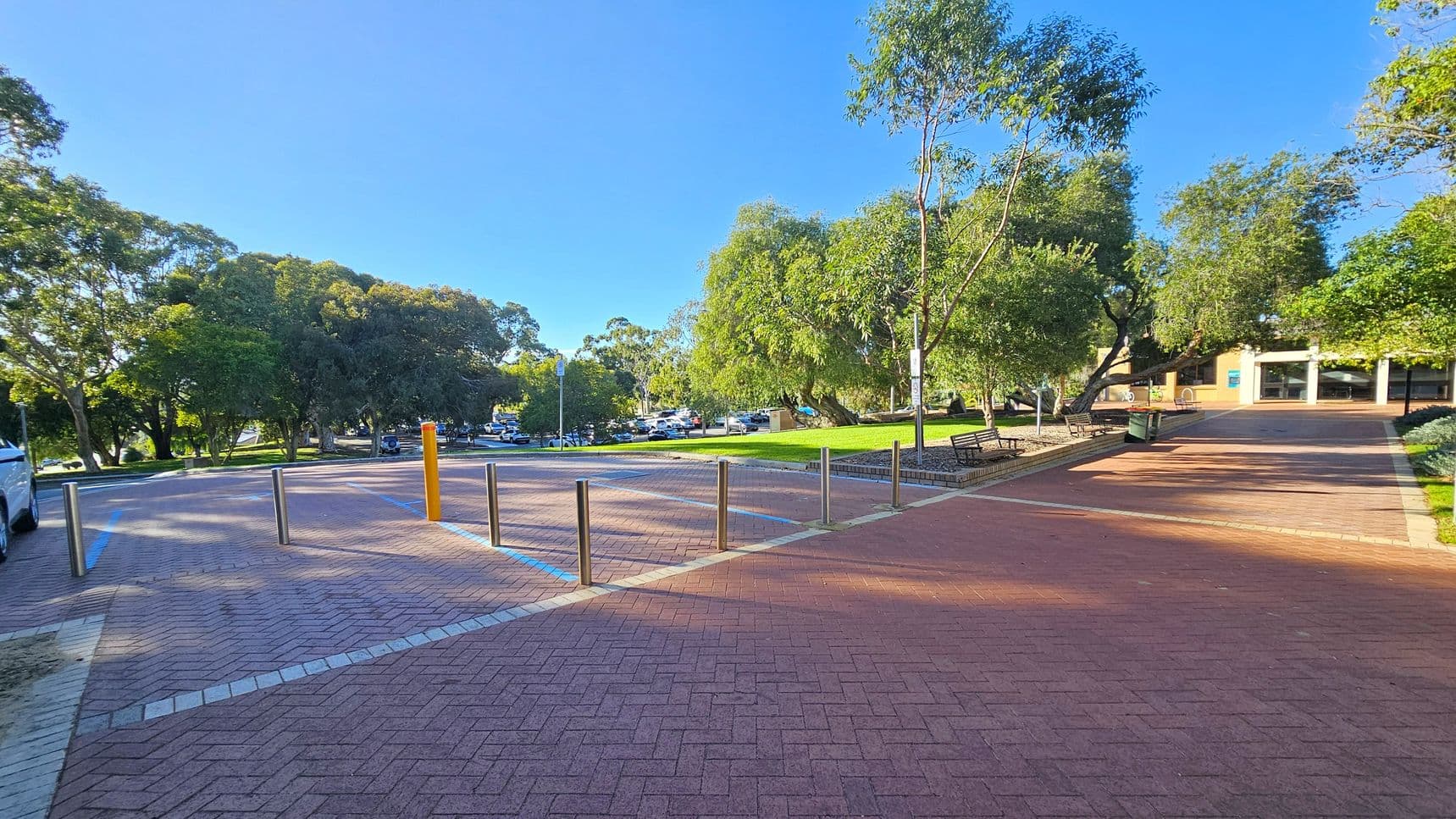 A paved area with a pattern of red bricks in the foreground, bordered by metal bollards. To the left, a row of parked cars is partially visible under the shade of large trees. In the centre, a grassy area with more trees and a few benches. On the right, a building with a light-coloured facade and large windows. The background features a parking lot with several cars and more trees. The scene is well-lit, suggesting a sunny day.