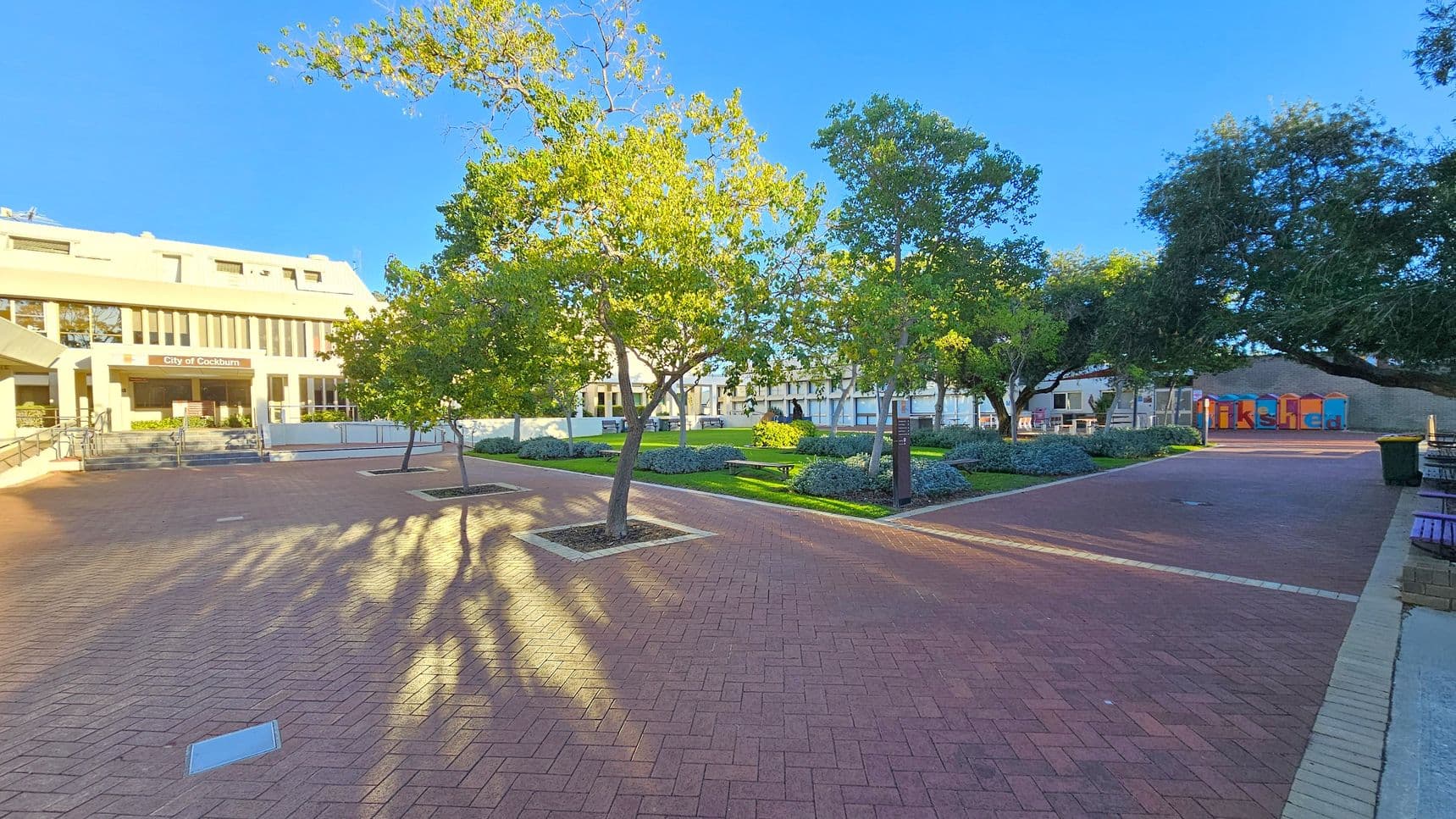 A paved courtyard with red bricks and several trees casting shadows. On the left, a building with a sign reading "City of Cockburn" and a series of steps leading up to it. In the centre, green grass and shrubs are neatly arranged. On the right, colourful structures with the word "Shed" visible, and a green bin nearby. Benches are placed along the edges of the courtyard.