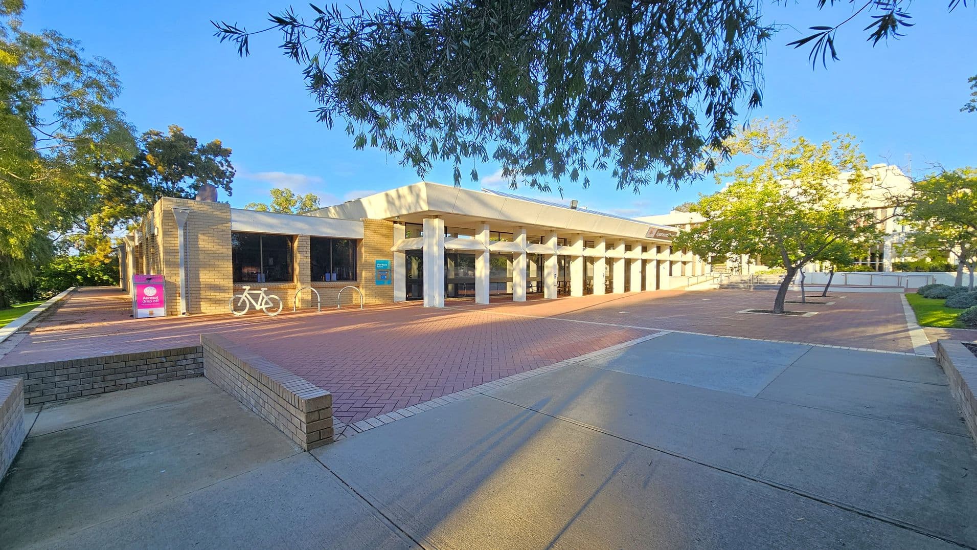 A building with a modern design featuring large windows and white columns is in the centre. To the left, there is a bicycle rack with a white bicycle and a pink sign. The foreground has a paved area with a mix of red and grey tiles. Trees with green foliage are on both sides, with branches extending over the scene. The background shows more trees and a clear blue sky.