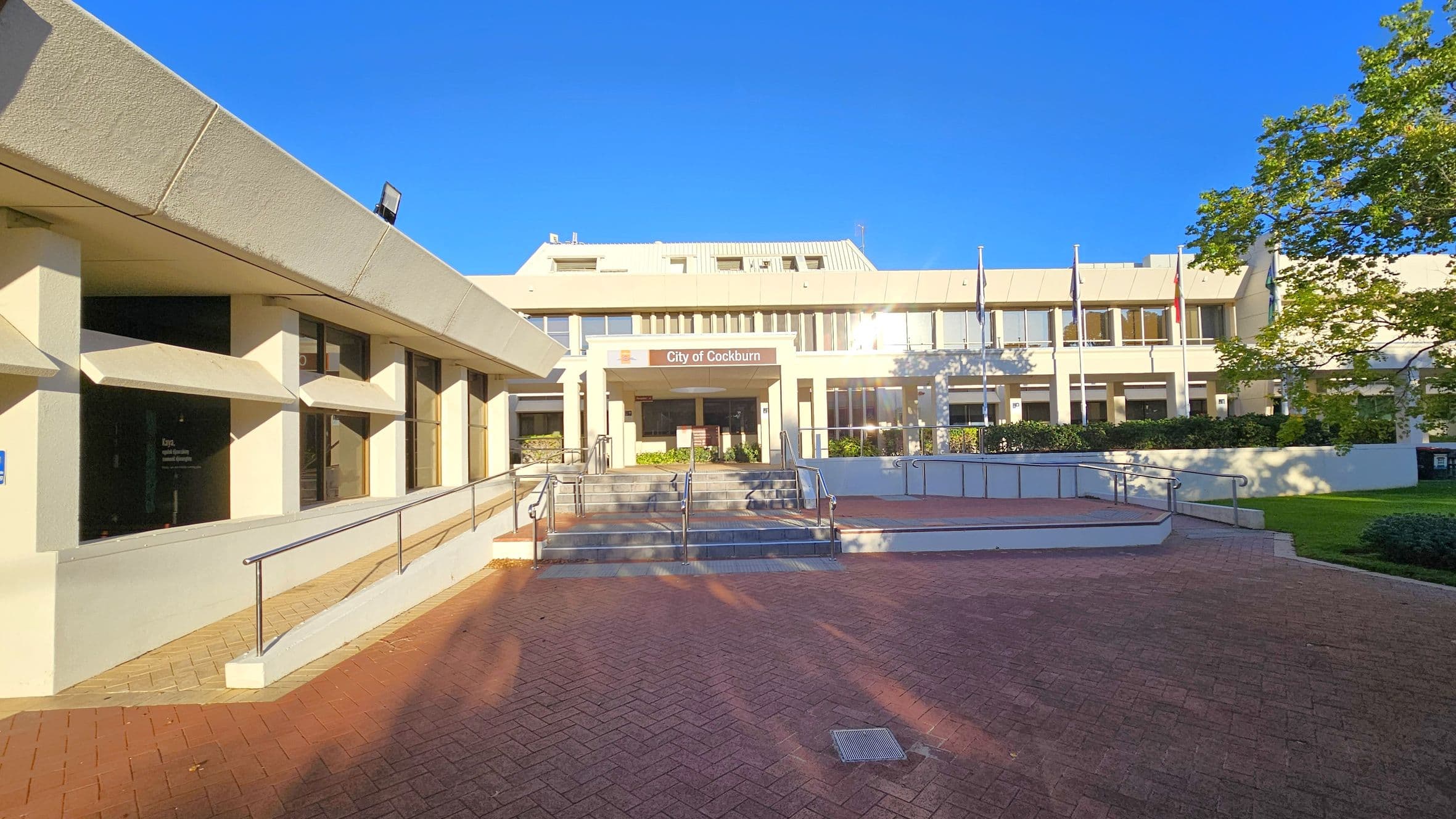A modern building with a beige facade and large windows is in the centre. The entrance has a sign above it. In front, there are steps with metal railings leading up to the entrance. On the left, a sloped walkway with a railing runs alongside the building. On the right, there are several flagpoles and a tree with green leaves. The foreground features a red brick paved area. The sky is clear and blue.