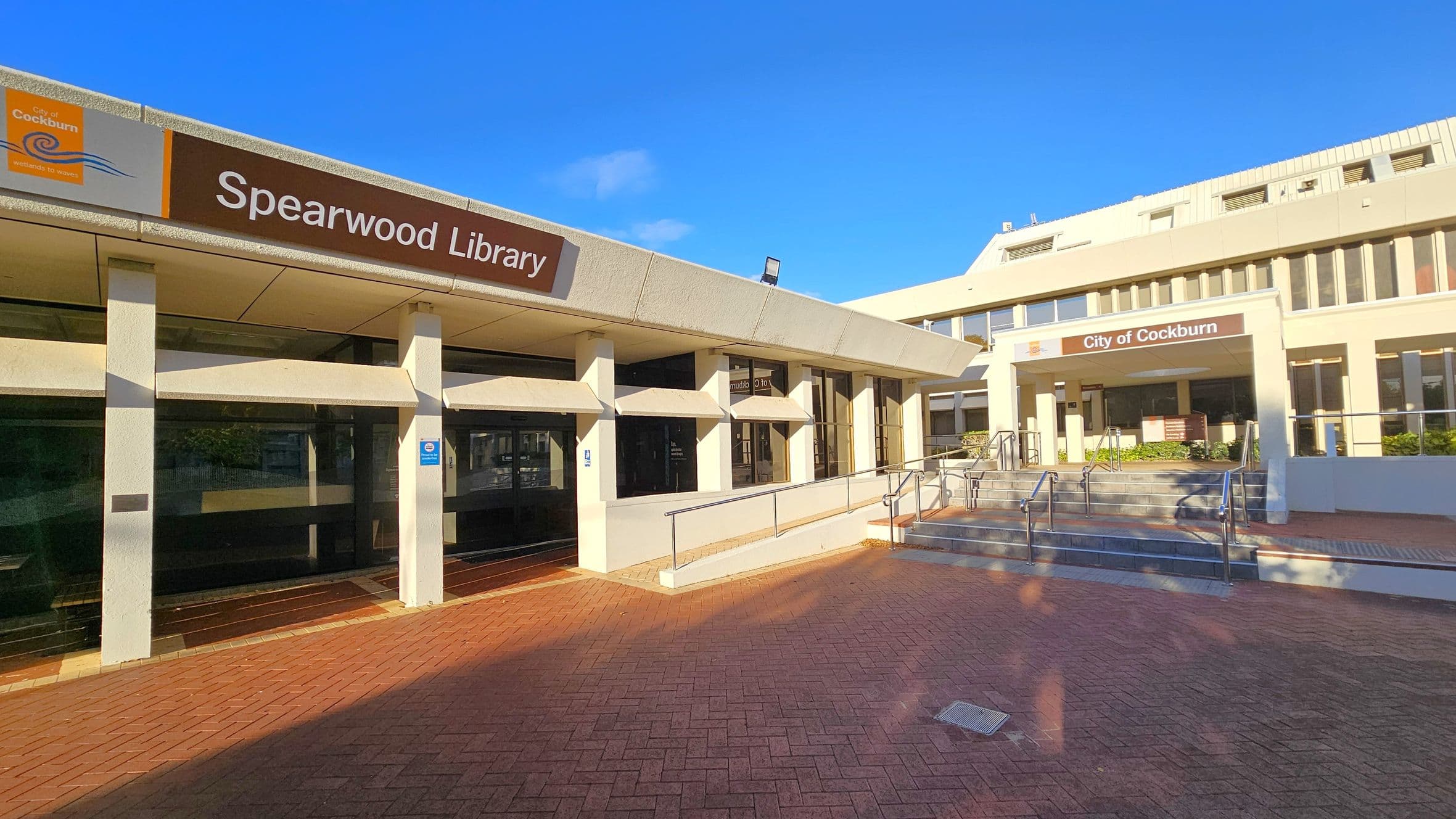 On the left, a building with a sign reading "Spearwood Library" and "City of Cockburn" is visible. The structure has large windows and a light-coloured facade. In the centre, a ramp with metal railings leads to the entrance. On the right, another section of the building with steps and additional railings is present. The ground is paved with red bricks, and the area is well-lit by sunlight.