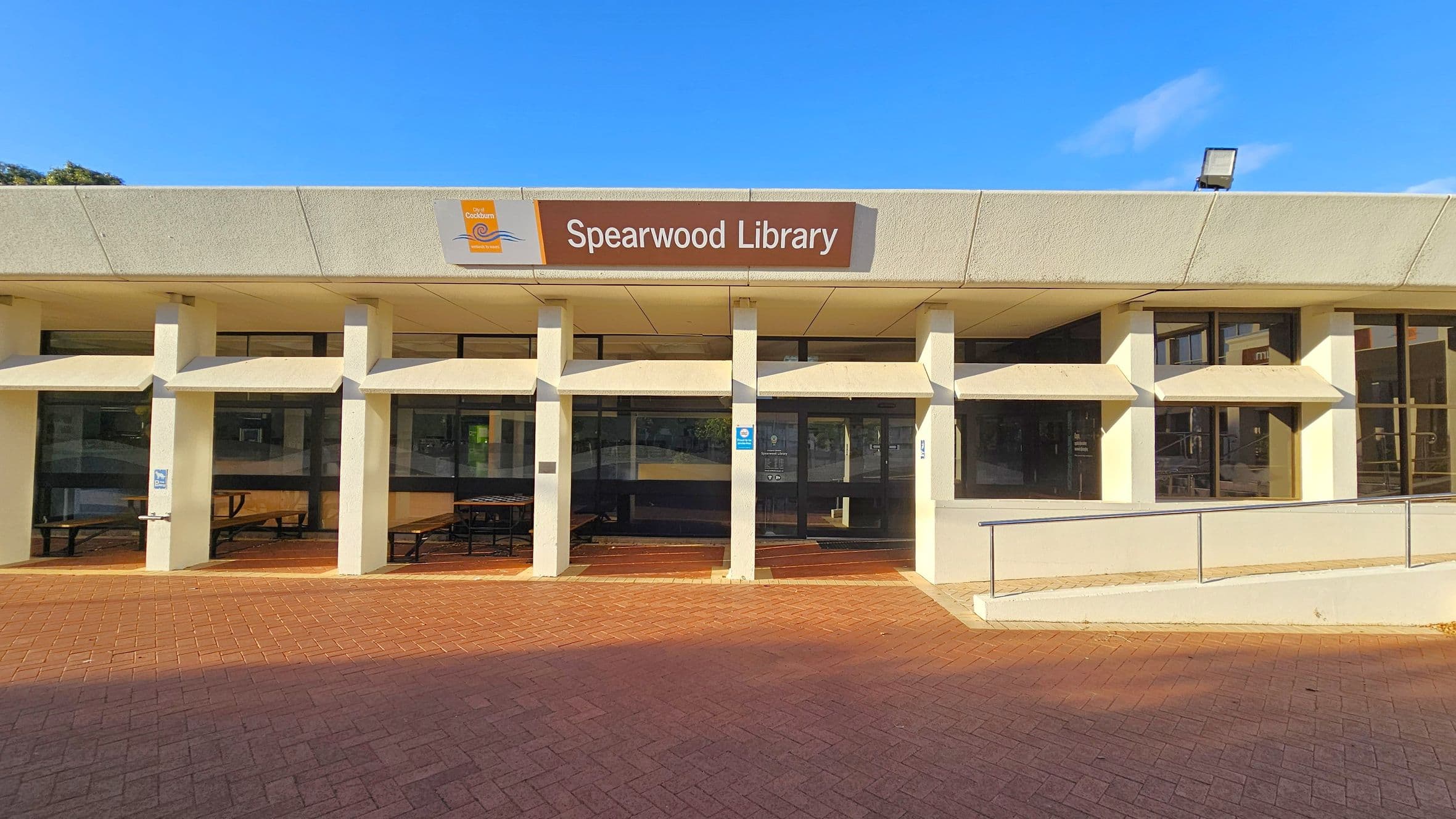 A building with a sign reading "Spearwood Library" is centred on the facade. The structure features a series of vertical white columns and large windows. The ground is paved with reddish-brown bricks, and there is a ramp with a metal railing on the right. The roof is flat with a light fixture visible on top.