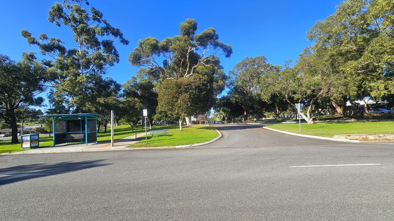 A road curves gently to the right, bordered by a grassy area with several large trees. On the left, there is a bus shelter with a bench inside, next to a rubbish bin and a signpost. A paved footpath runs parallel to the road. The area is well-shaded by the trees, and the grass is neatly trimmed. In the background, more trees and a few parked cars are visible. The sky is clear and blue.