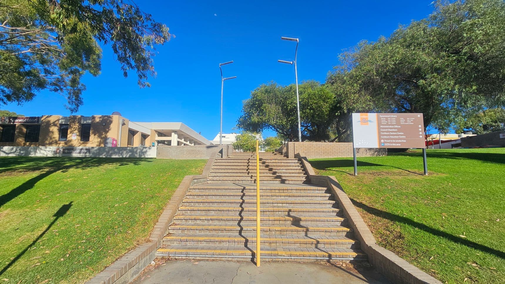Wide stone steps with a yellow handrail in the centre lead up to a brick building on the left, labelled "Spearwood Library". A sign on the right lists various facilities. Two tall street lamps are positioned at the top of the steps. The area is surrounded by green grass and trees, with a clear blue sky above.