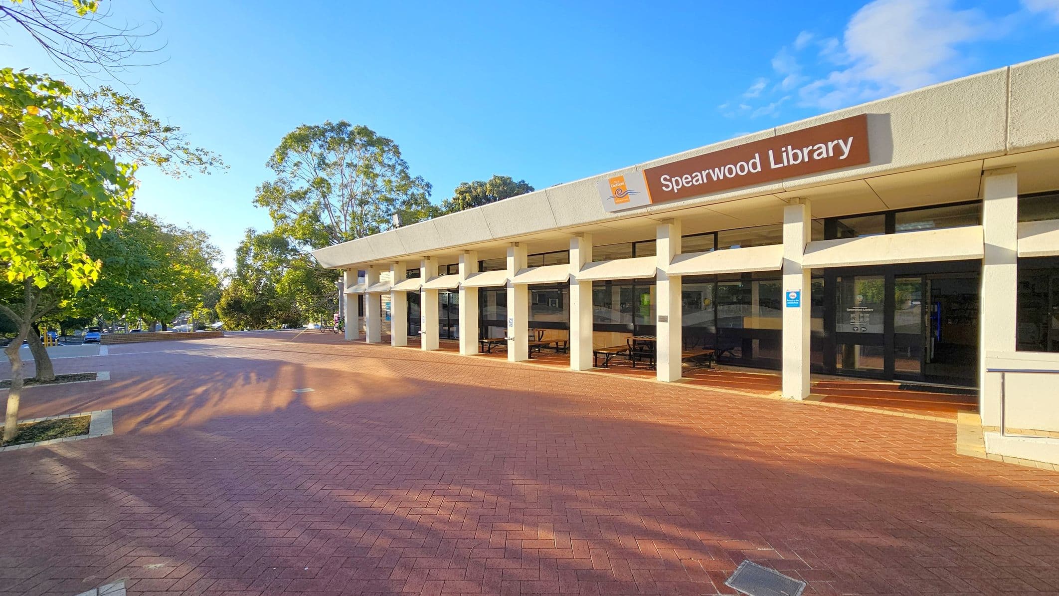 A modern building with a flat roof and large windows on the right, labelled "Spearwood Library". The structure is supported by white columns. The foreground features a red brick paved area, casting shadows from nearby trees. On the left, there are several green trees and a few parked bicycles. The background includes more trees and a clear blue sky.