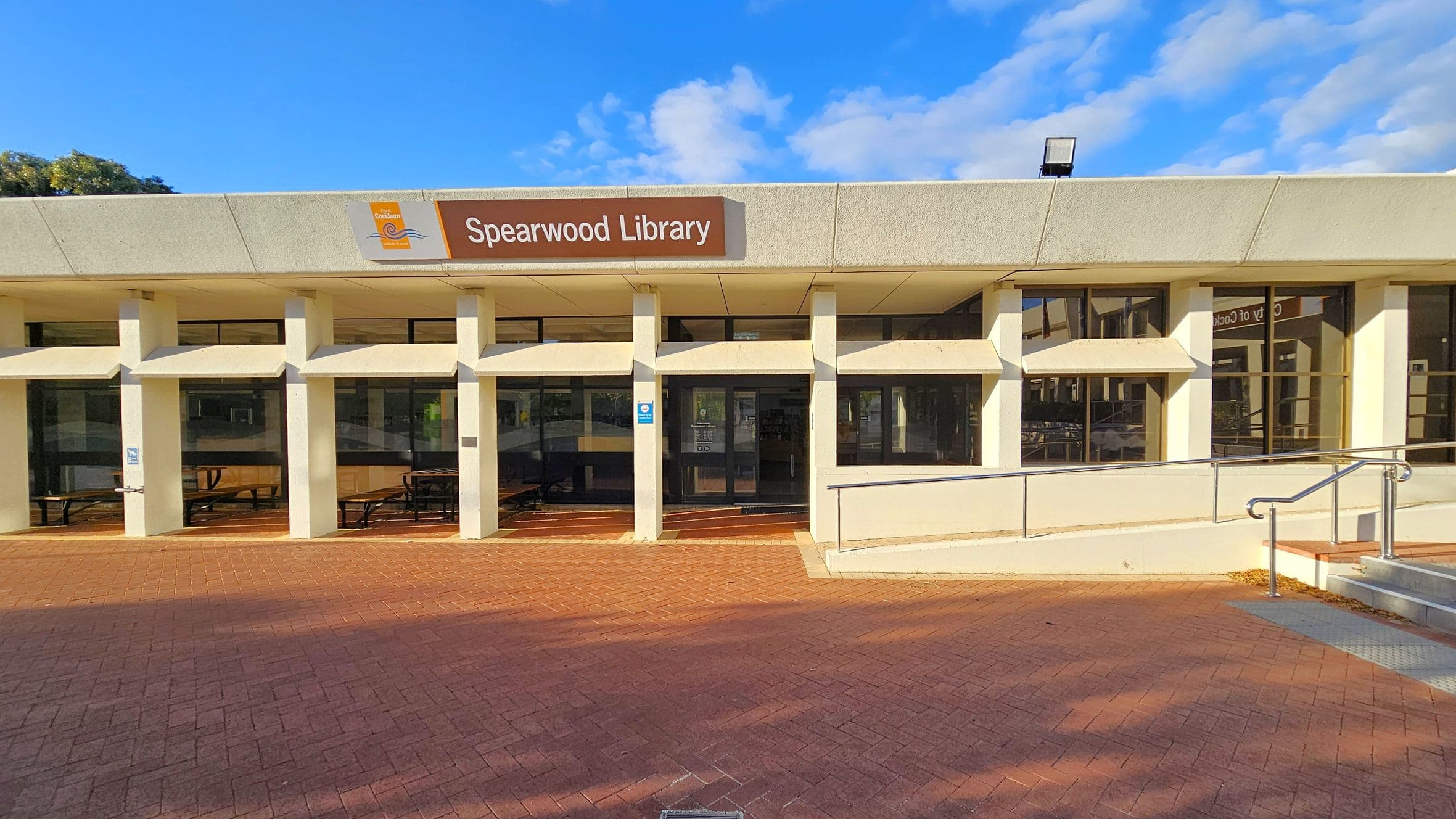 A modern building with a flat roof and a sign reading "Spearwood Library" above the entrance. The facade features large windows and white pillars. The ground is paved with red bricks, and there is a ramp with metal railings on the right side. The entrance has glass doors, and there are picnic tables visible inside through the windows.