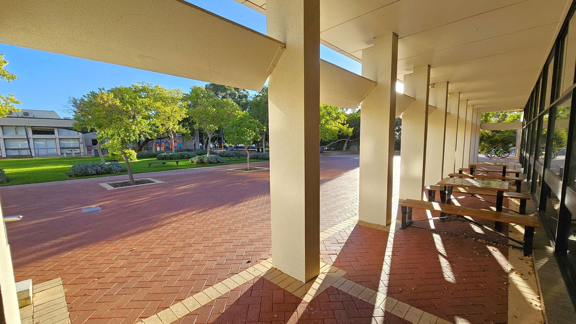 A covered walkway with a series of vertical columns on the right, casting shadows on the red brick floor. Wooden benches are placed along the right side, adjacent to a building with large windows. On the left, there is an open area with green grass, trees, and a distant building. The ceiling is flat and light-coloured, with beams extending from the columns.
