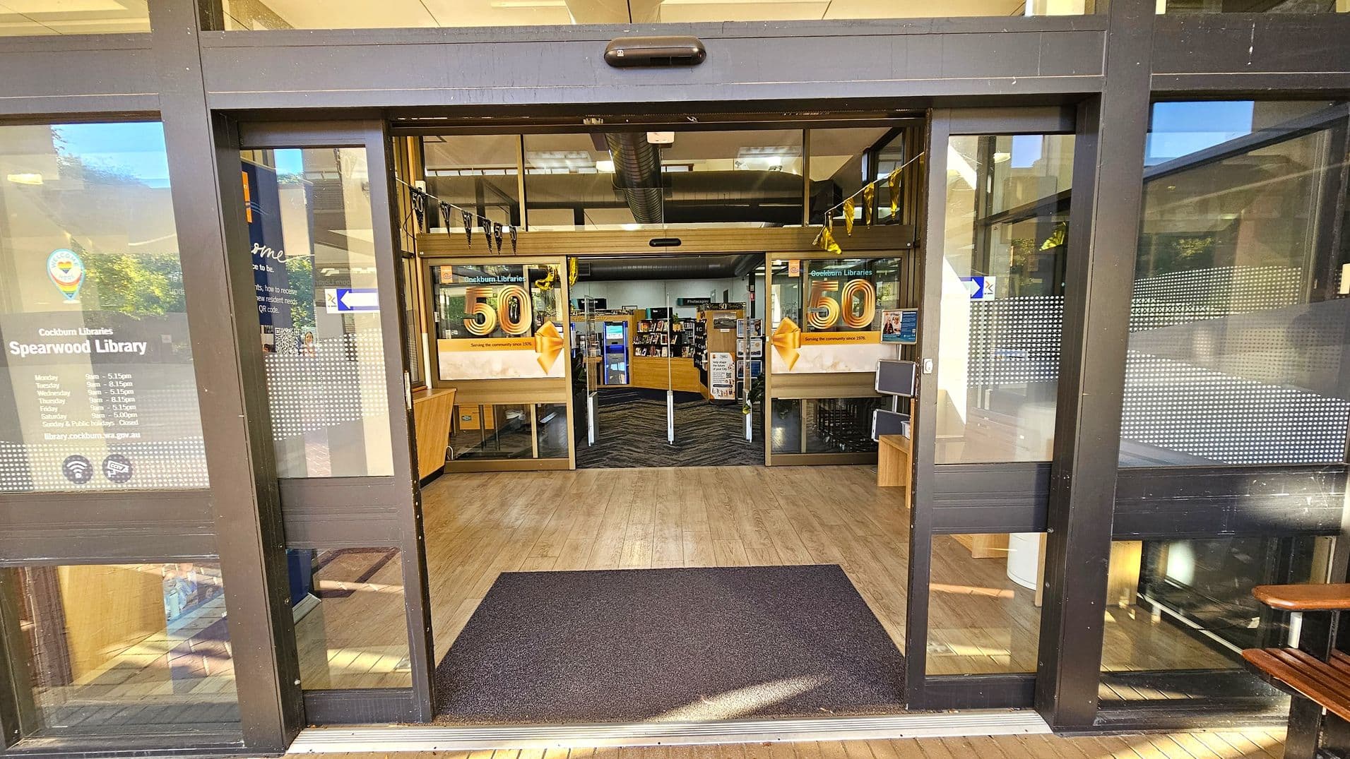 Entrance to a library with glass sliding doors. On the left door, there's information about the library's opening hours. Inside, a banner celebrates the library's 50th anniversary. The floor is wooden with a dark mat at the entrance. Inside, bookshelves and a counter are visible. The ceiling has exposed ductwork and lighting fixtures.