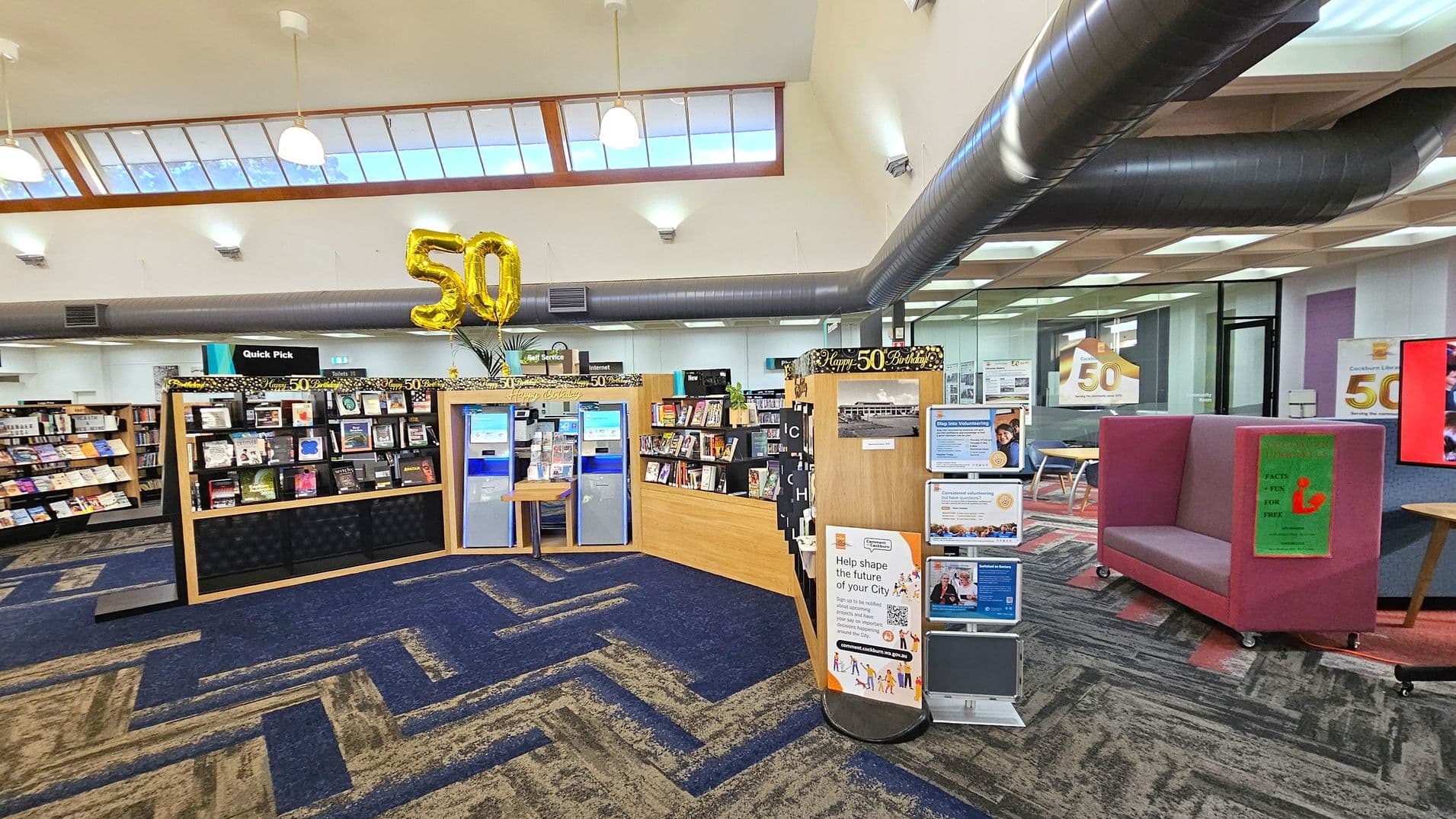 A library interior with a blue and grey patterned carpet. In the centre, a display with books and two self-service machines is decorated with gold "50" balloons. To the right, a pink cushioned seating area is visible, along with a digital screen. The ceiling has skylights and hanging lights, and there are large air ducts. Bookshelves are on the left, and a glass-walled room is in the background. Signs and posters are displayed throughout the space.