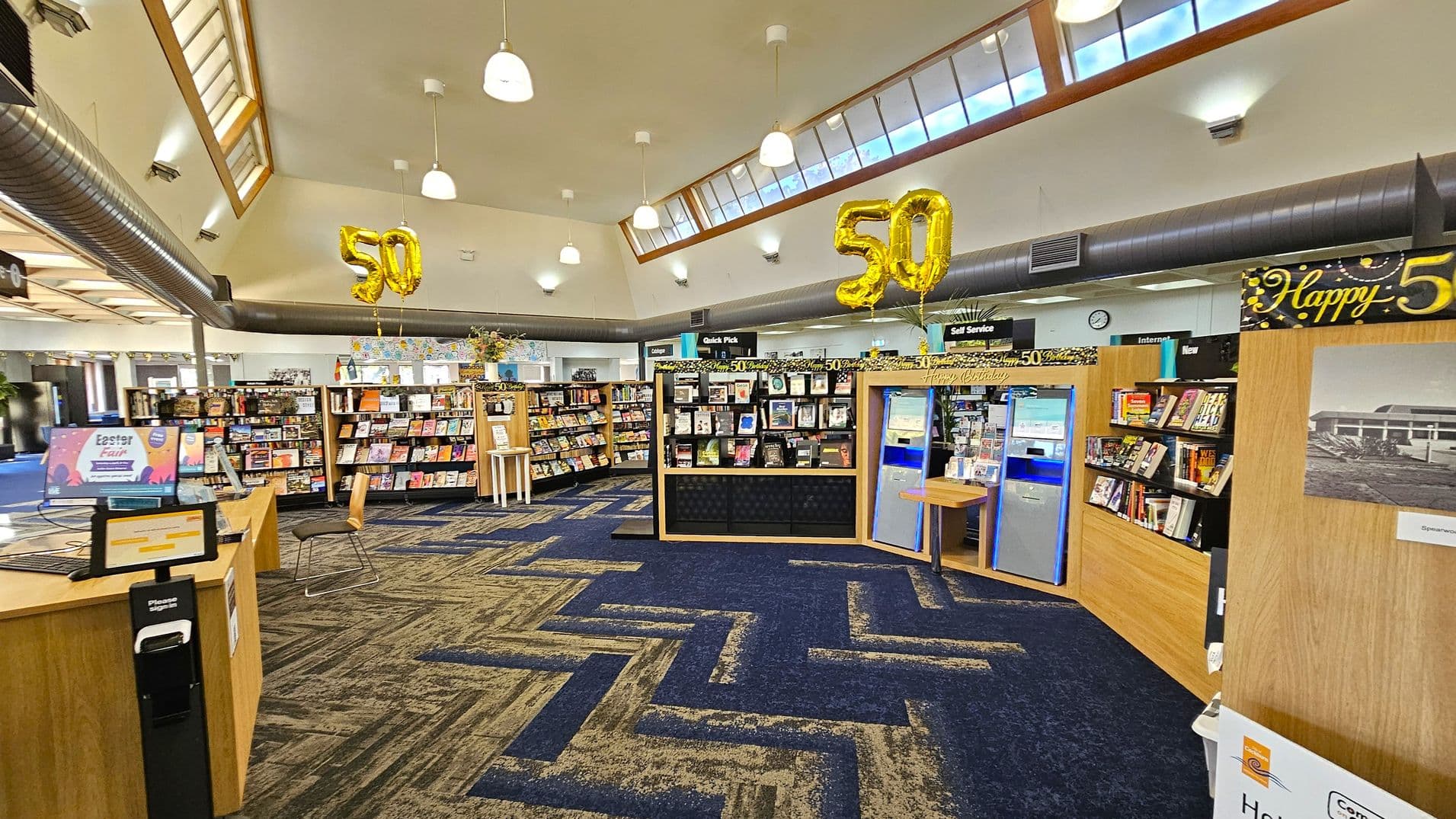 A library interior with a high ceiling and large windows. In the centre, there are bookshelves filled with books and magazines. Golden balloons shaped as the number "50" are suspended from the ceiling, indicating a celebration. The floor has a patterned carpet in shades of blue and grey. On the right, there are self-service machines and a display with a "Happy 50" sign. The left side features a computer station and a chair. Hanging lights illuminate the space.