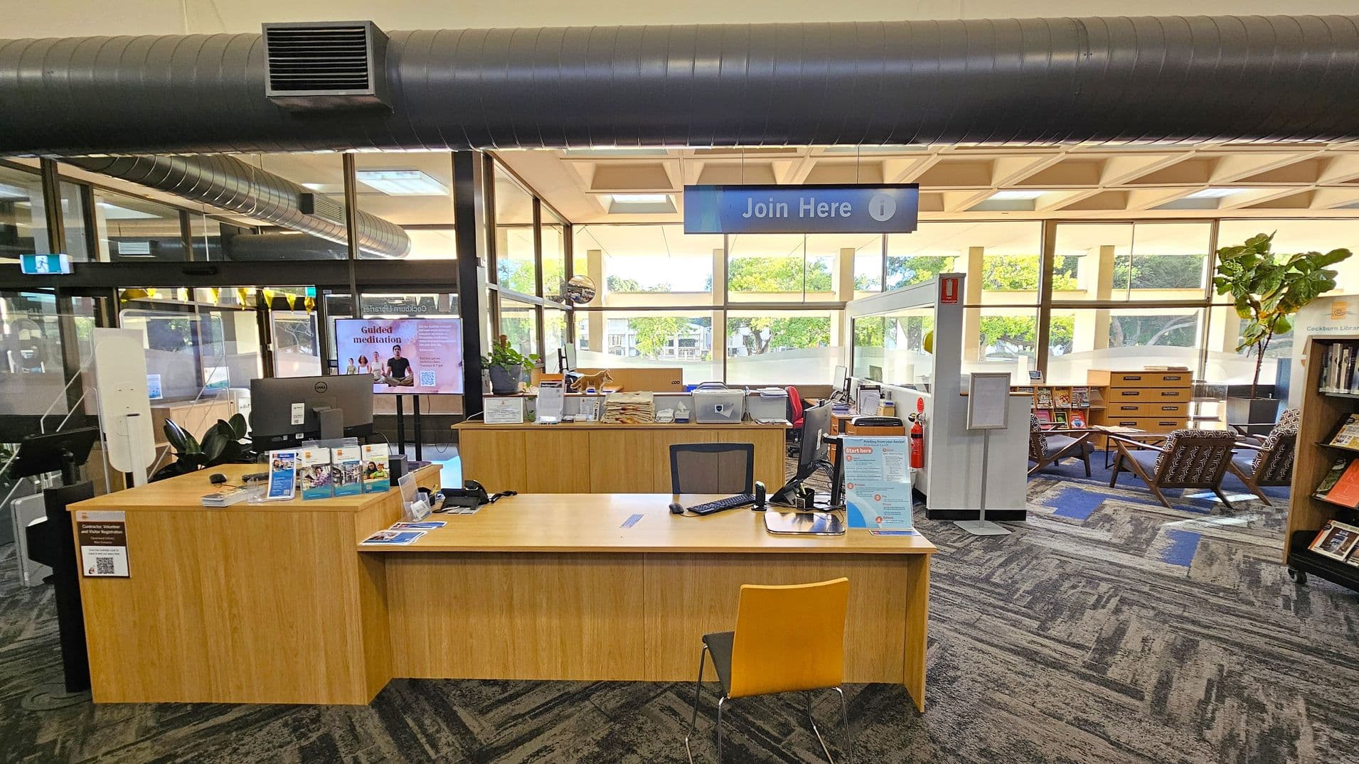 A library interior with a wooden reception desk in the foreground, featuring a computer, brochures, and a yellow chair. Above, a sign reads "Join Here". The background has large windows allowing natural light, with a view of trees outside. To the right, there are bookshelves and a seating area with patterned chairs and a small table. The ceiling has exposed ductwork, and the floor is covered with a patterned carpet.
