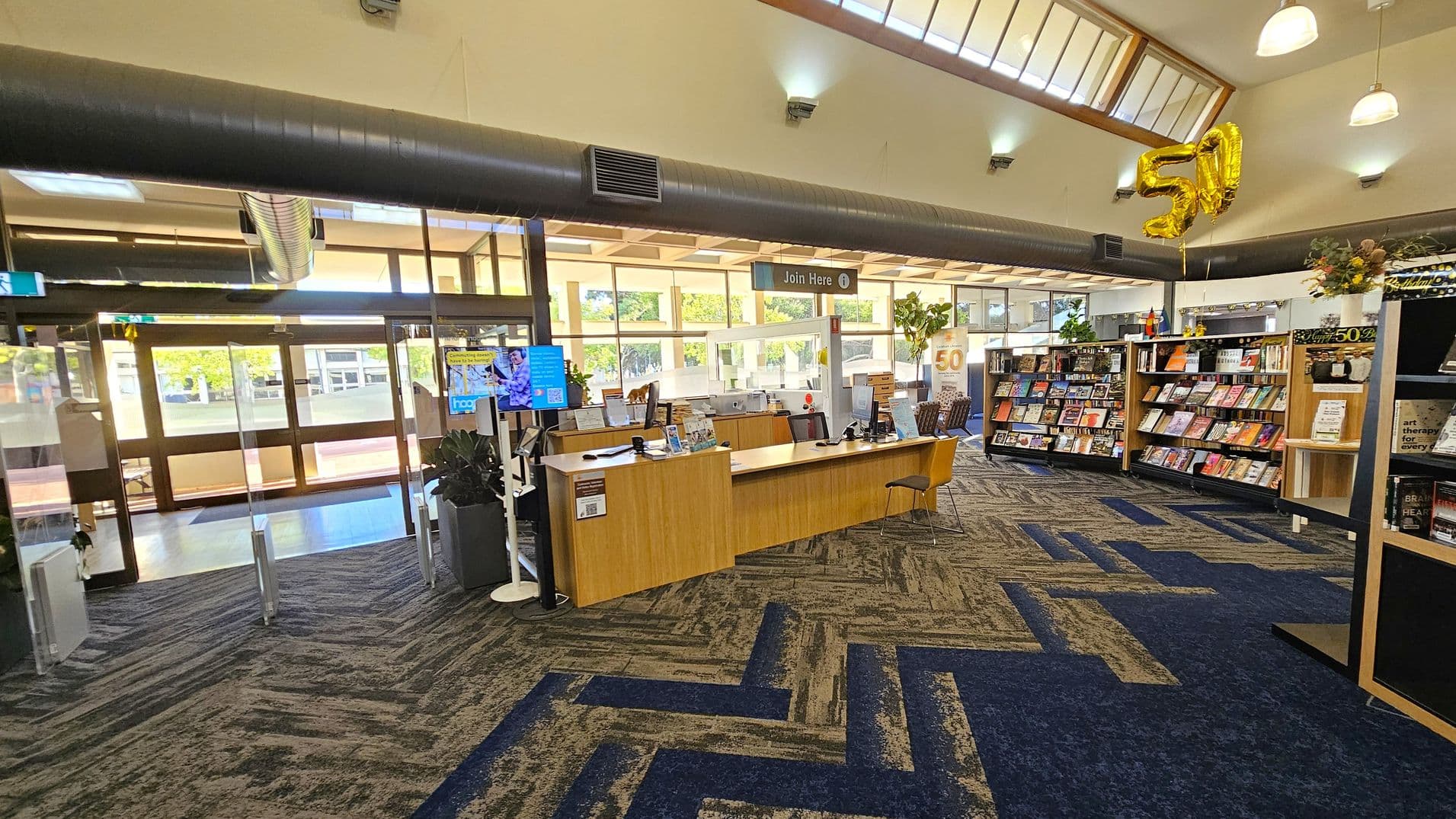A library interior with a patterned carpet in shades of blue and brown. In the centre, a wooden reception desk with a "Join Here" sign above it. To the right, bookshelves filled with books and a golden "50" balloon. The ceiling has skylights and hanging lights. Large windows in the background allow natural light to enter, and there are plants near the entrance.