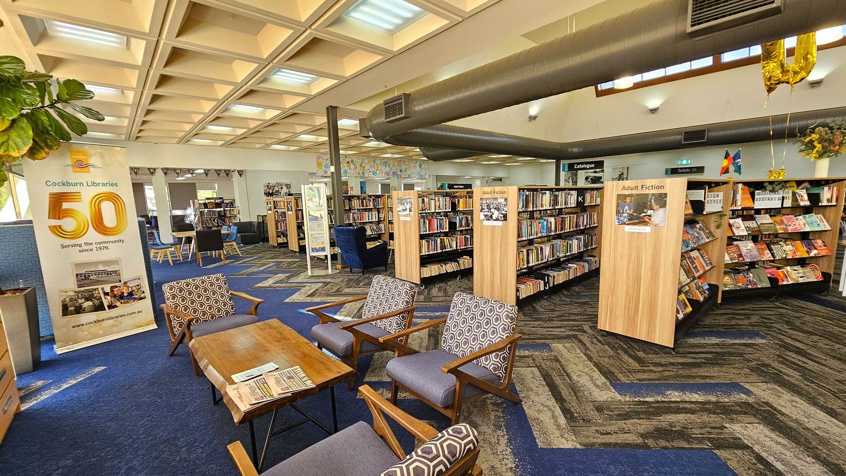 A library interior with a blue and patterned carpet. On the left, a banner celebrates "50 years" of Cockburn Libraries. In the foreground, there are wooden chairs with patterned cushions and a wooden table with newspapers. The middle features bookshelves labelled "Adult Fiction" filled with books. The ceiling has a grid pattern with skylights, and a large air duct runs across. In the background, there are more bookshelves and a sign for the catalogue.