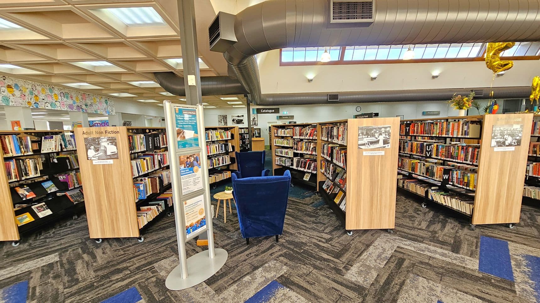 A library interior with bookshelves filled with books on both sides. In the centre, there are two blue armchairs and a small round table. A stand with informational posters is positioned in front of the chairs. The ceiling features a grid pattern with fluorescent lights and large air ducts. The floor is carpeted with a geometric pattern in grey and blue tones. Signs for "Adult Non Fiction" and "Catalogue" are visible, along with decorative balloons and artwork on the walls.