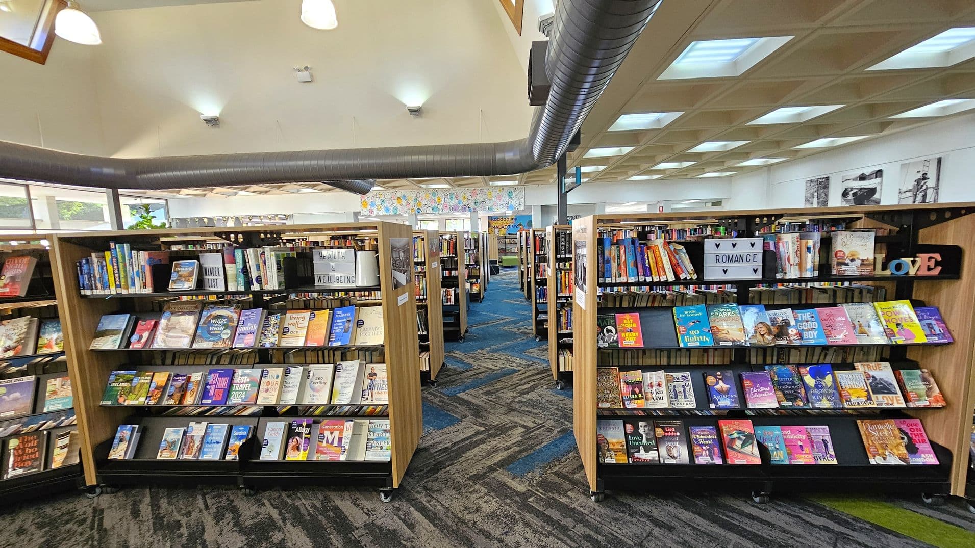 Bookshelves filled with various books are arranged in rows. The left shelf features books on health and well-being, while the right shelf displays romance novels. The floor is carpeted with a blue and green pattern. The ceiling has a grid design with recessed lighting and visible air ducts. In the background, there are more bookshelves and a colourful mural on the wall.