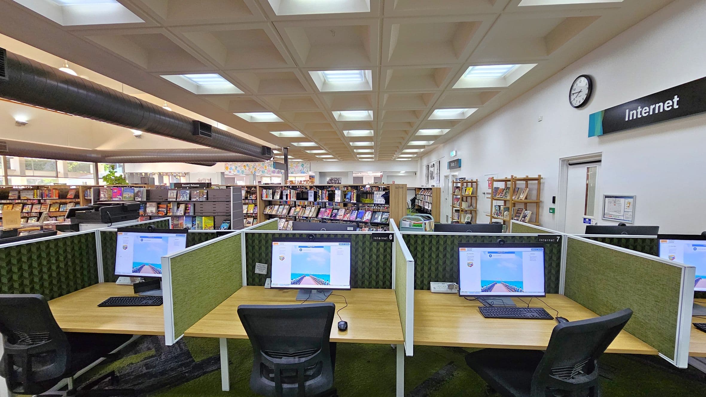 A library interior with computer workstations in the foreground, each equipped with a monitor, keyboard, and mouse on wooden desks. Green fabric partitions separate the stations. In the background, bookshelves filled with books are visible. The ceiling has a grid pattern with recessed lighting, and a large black duct runs across it. A clock and a sign reading "Internet" are mounted on the right wall. The floor is carpeted in a dark pattern.
