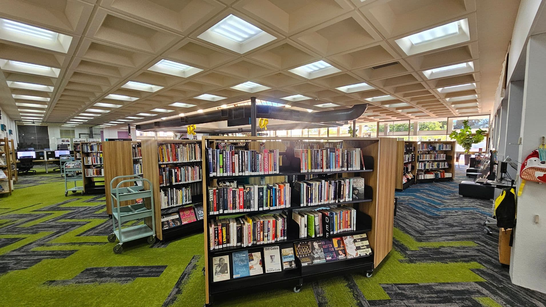 Bookshelves filled with books are in the centre of a library. On the left, there are computer stations and a green book cart. The floor has a green and grey zigzag pattern. On the right, there are more bookshelves and a computer station with a chair. The ceiling features a grid pattern with recessed lighting. Large windows in the background allow natural light to enter, and there are plants visible outside.