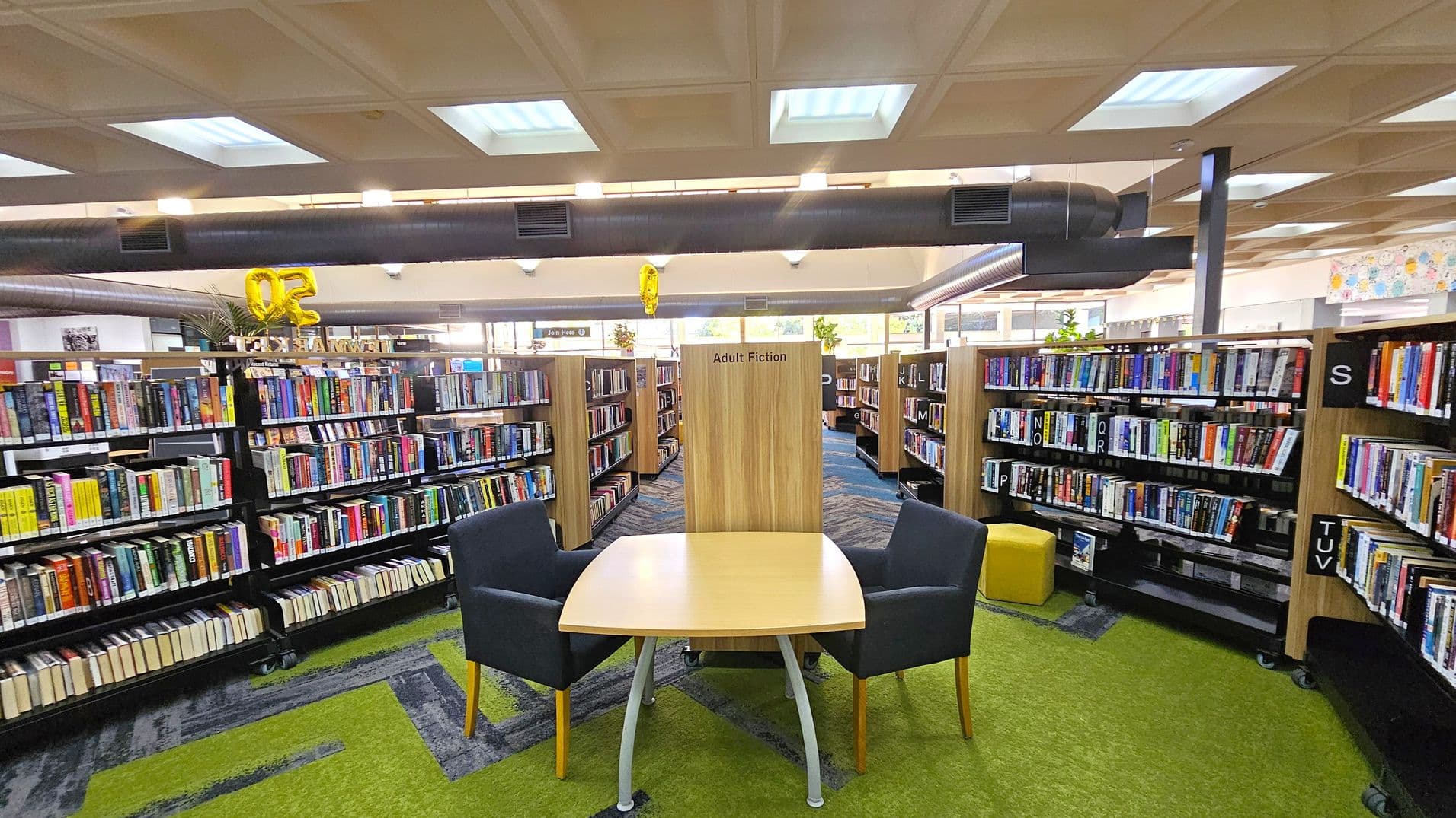 A library interior with a focus on the "Adult Fiction" section. In the foreground, a wooden table with two dark chairs is placed on a green carpet. Bookshelves filled with books are arranged in rows on both sides. A yellow balloon with the number "02" is visible above the shelves. The ceiling has a grid pattern with fluorescent lights, and a large air duct runs across it. The floor is a mix of green and blue carpeting.