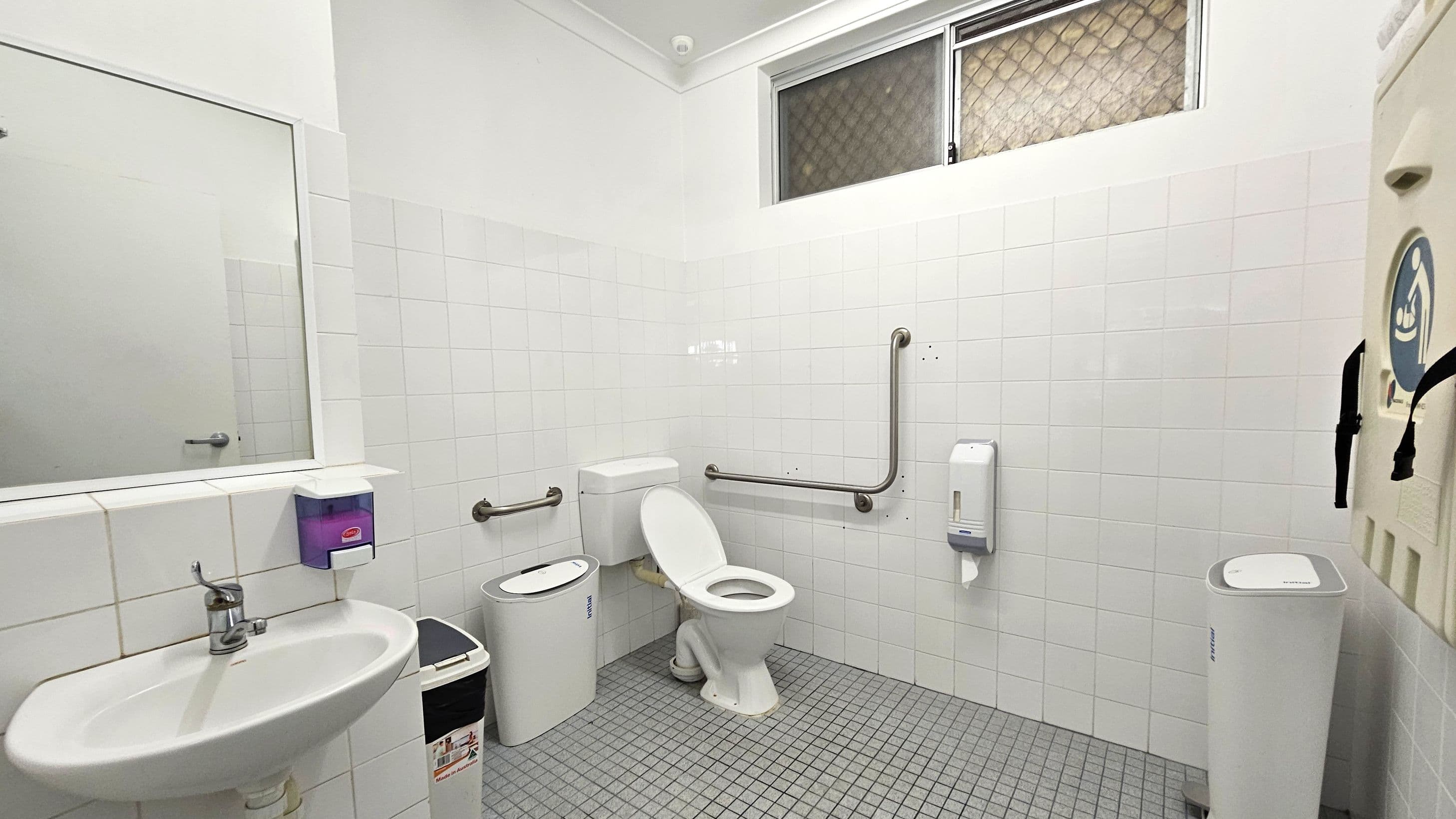 A public restroom with white tiled walls and a grey tiled floor. On the left, a sink with a mirror above it and a soap dispenser mounted on the wall. In the centre, a toilet with grab bars on both sides for accessibility. To the right, a nappy changing station is mounted on the wall, and a paper towel dispenser is nearby. A small window with a mesh screen is located high on the wall.