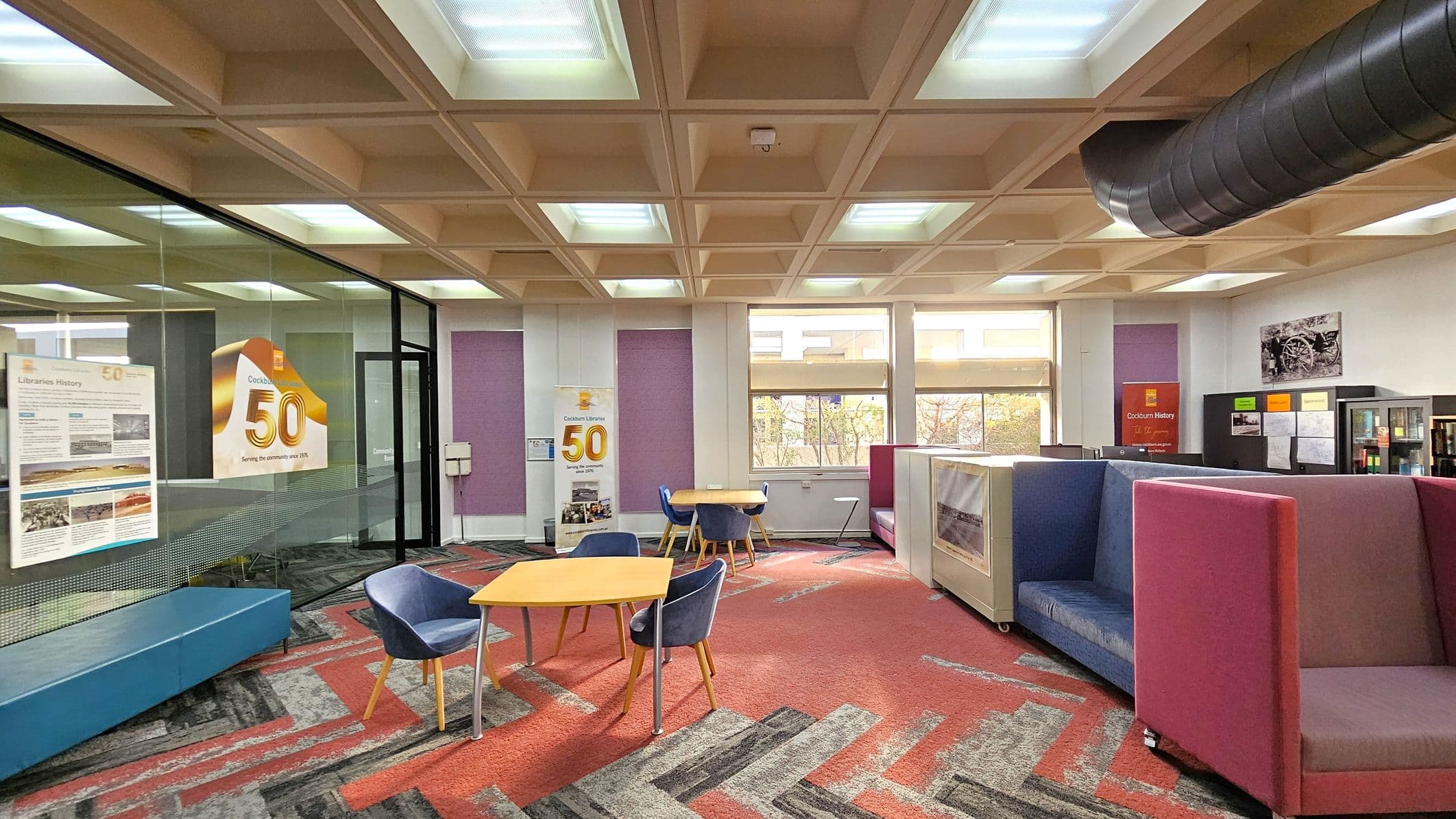 A modern library interior with a patterned carpet in red and grey tones. On the left, a glass wall displays posters, including one celebrating a 50th anniversary. In the centre, there are tables with blue chairs. The right side features colourful, high-backed seating booths. Large windows in the background allow natural light in. The ceiling has a grid pattern with recessed lighting, and a black ventilation duct runs across it.