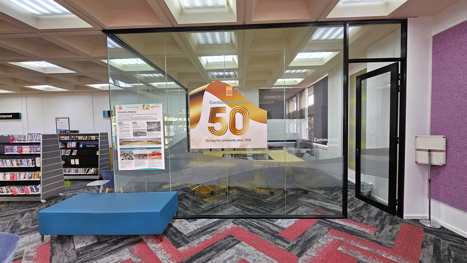 A library interior with a glass-walled community room in the centre. The glass displays a large sign celebrating "50" years of service. To the left, there are bookshelves filled with DVDs and books, and a blue bench. The floor has a patterned carpet in grey and red. The ceiling features recessed lighting panels. On the right, a purple wall and a small white box are visible.