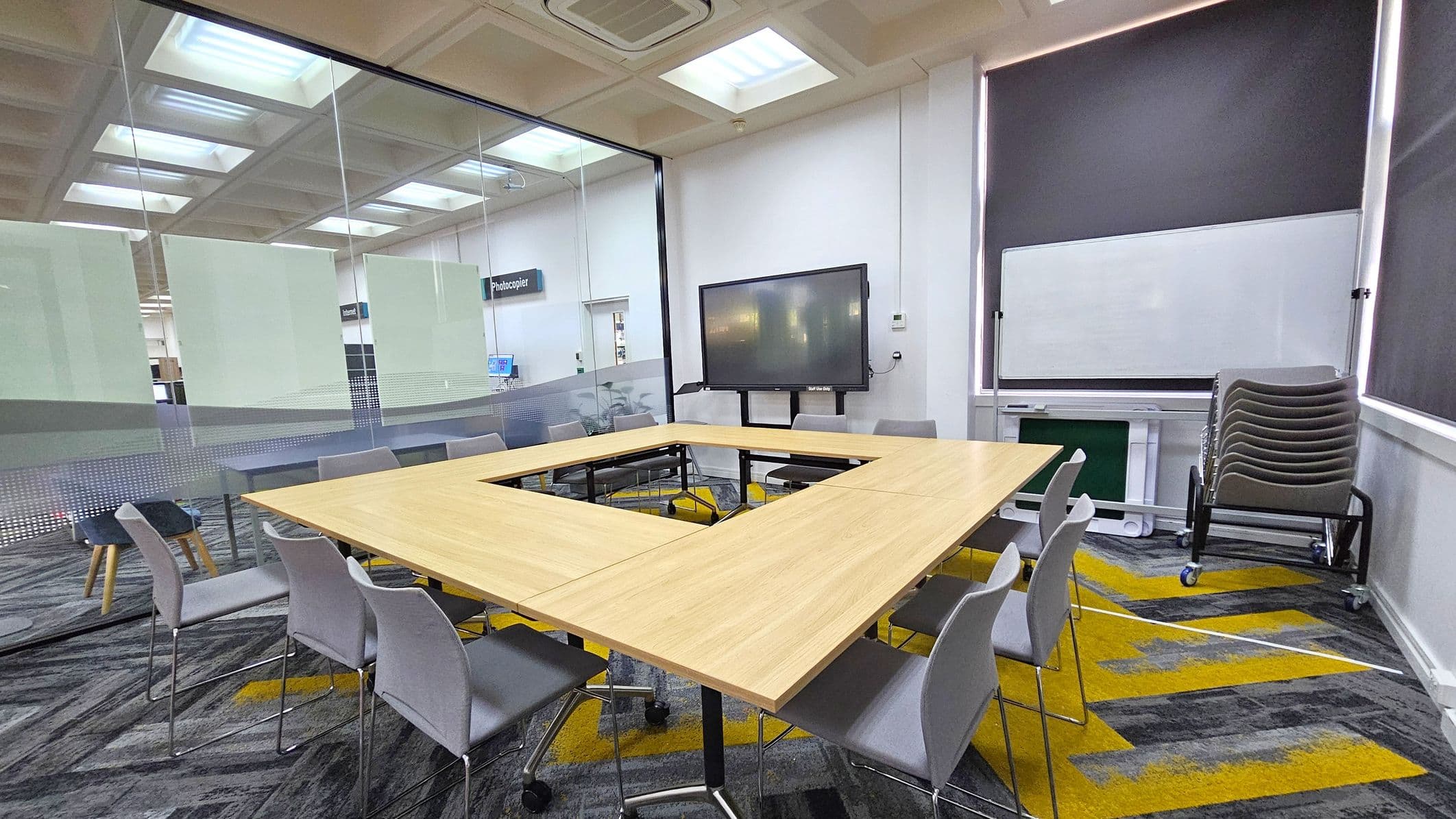 A meeting room with a U-shaped wooden table surrounded by grey chairs. On the right, a stack of additional chairs is placed near a whiteboard. A large screen is mounted on the wall. The floor has a patterned carpet with grey and yellow designs. The left side features a glass wall with frosted panels, revealing a photocopier area outside. The ceiling has recessed lighting and air conditioning vents.