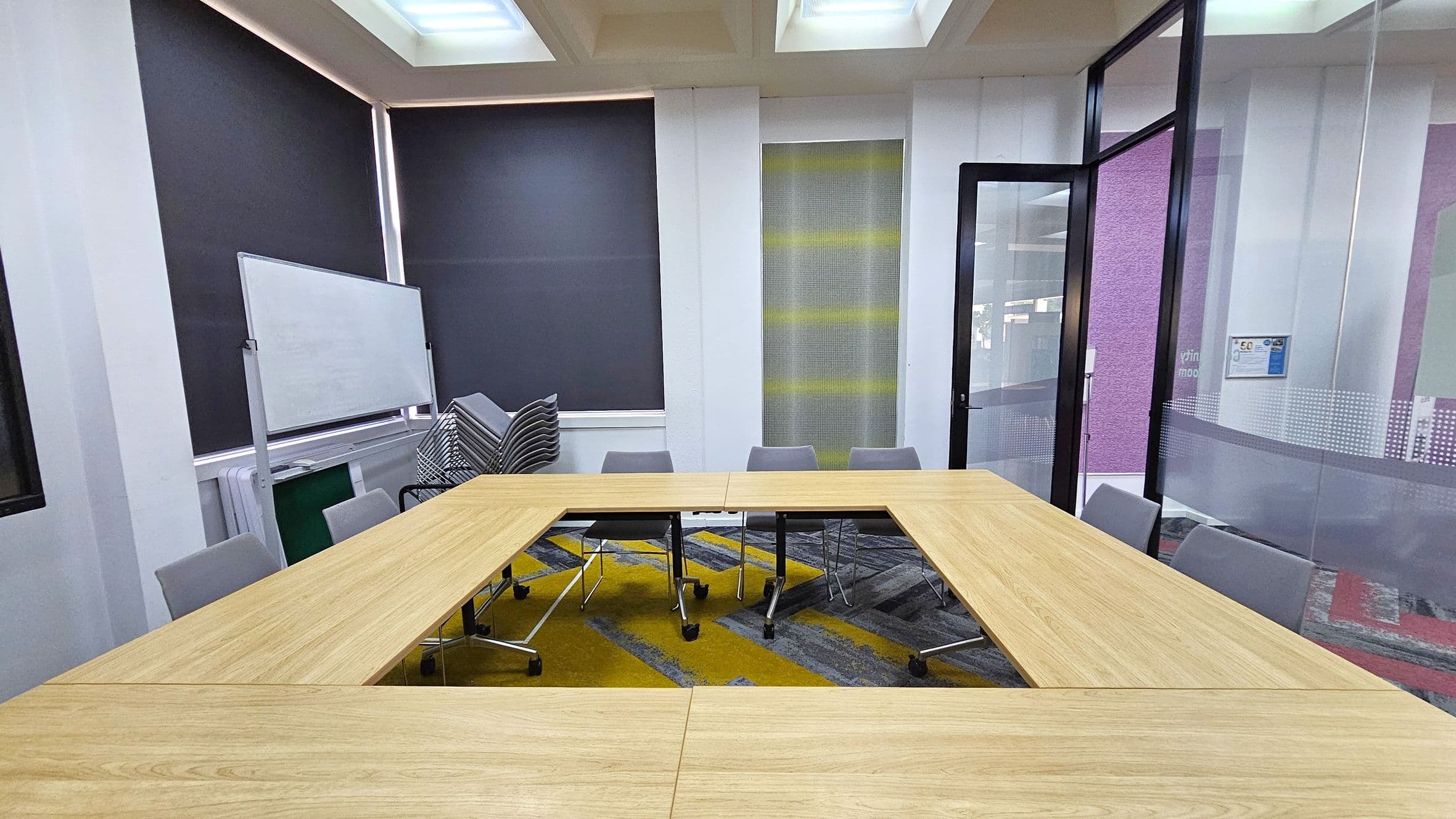 A meeting room with a U-shaped wooden table surrounded by grey chairs. On the left, a whiteboard is positioned against the wall, with a stack of chairs nearby. The walls have large windows covered with dark blinds. On the right, a glass door is open, leading to another area. The floor is carpeted with a geometric pattern in grey and yellow. The ceiling features recessed lighting panels.