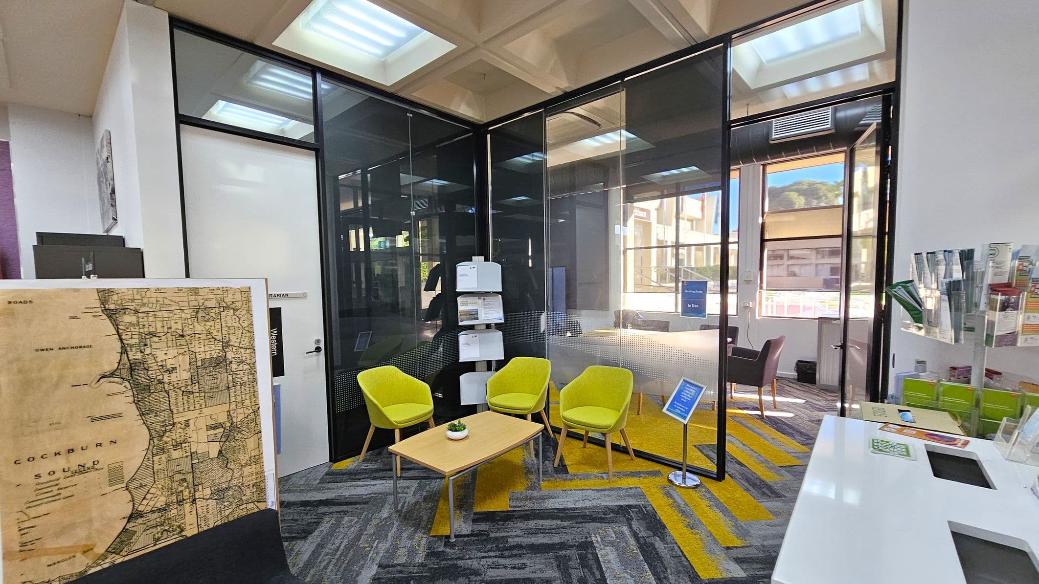 A modern office space with a glass-walled meeting room in the centre. Inside, there are three bright green chairs around a light wooden table with a small plant. To the left, a large vintage map is displayed. On the right, a stand holds brochures and pamphlets. The floor features a patterned carpet in grey and yellow. The ceiling has recessed lighting, and large windows allow natural light to enter, offering a view of the outside.