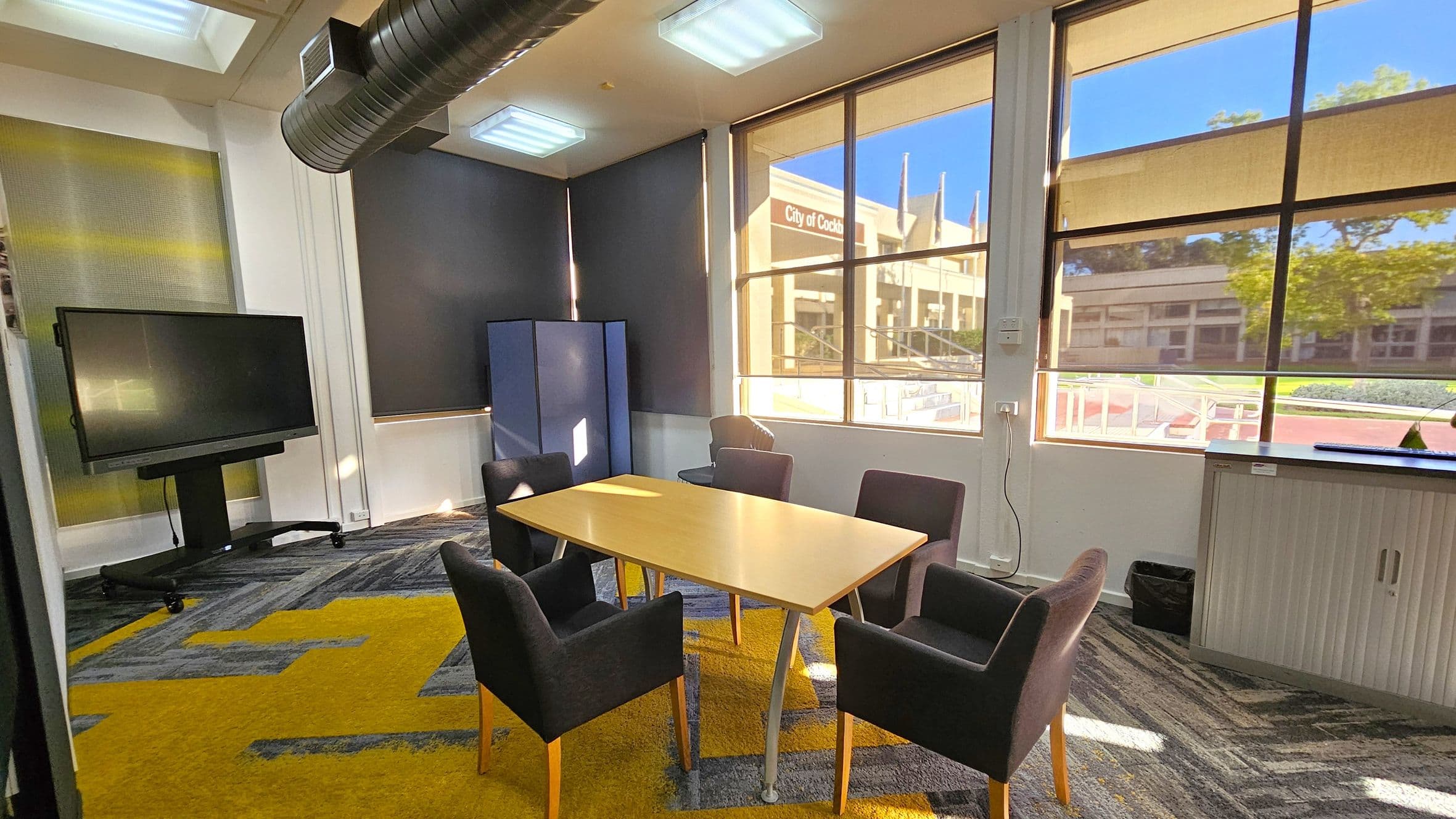 A meeting room with a rectangular wooden table surrounded by five dark upholstered chairs. On the left, a large screen on a stand is positioned near a wall with a textured panel. The floor has a patterned carpet in grey and yellow. Large windows on the right let in natural light, revealing an exterior view of a building and greenery. The ceiling has rectangular light fixtures and a visible air duct. A cabinet is against the wall beneath the windows.