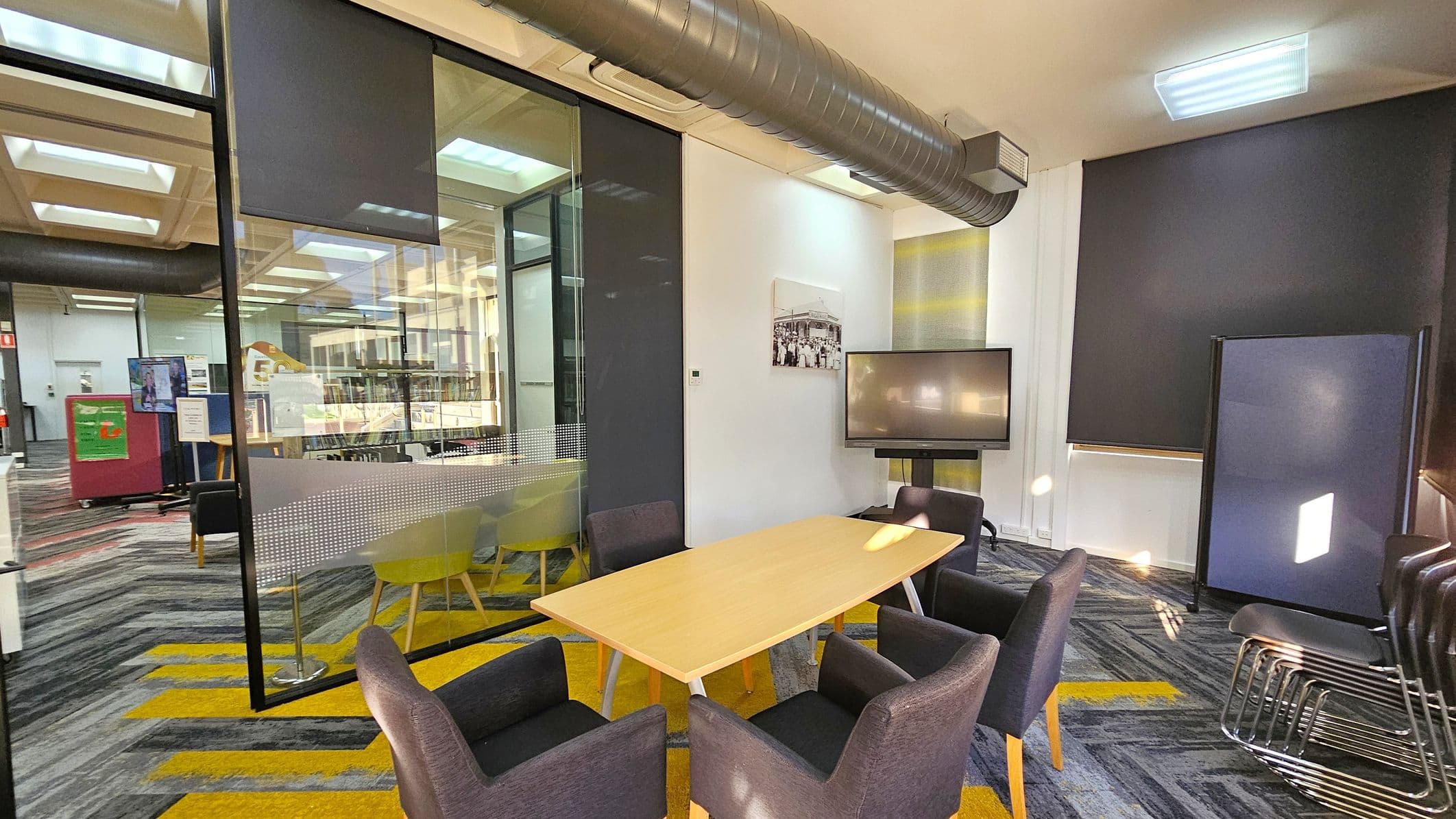 A modern meeting room with a light wooden table surrounded by six dark grey chairs. To the right, a large screen is mounted on the wall, and a stack of chairs is nearby. The floor has a patterned carpet with grey and yellow stripes. The ceiling features exposed ductwork and recessed lighting. On the left, a glass partition separates the room from an open area with more seating and notice boards. A black roller blind covers the window on the right wall.