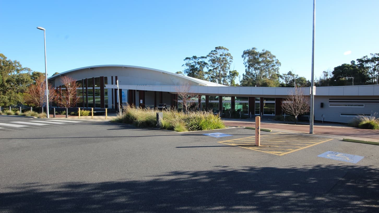 There is a pedestrian lane leading toward a fully accessible entrance-style ramp in Splash Devonport, and on the right is a plant box containing several bushes with moderate leaf length. Street lamp posts are scattered in the accessible parking spaces, distinguished by the persons with blue and white accessible parking signs, yellow bollards, and yellow lines.