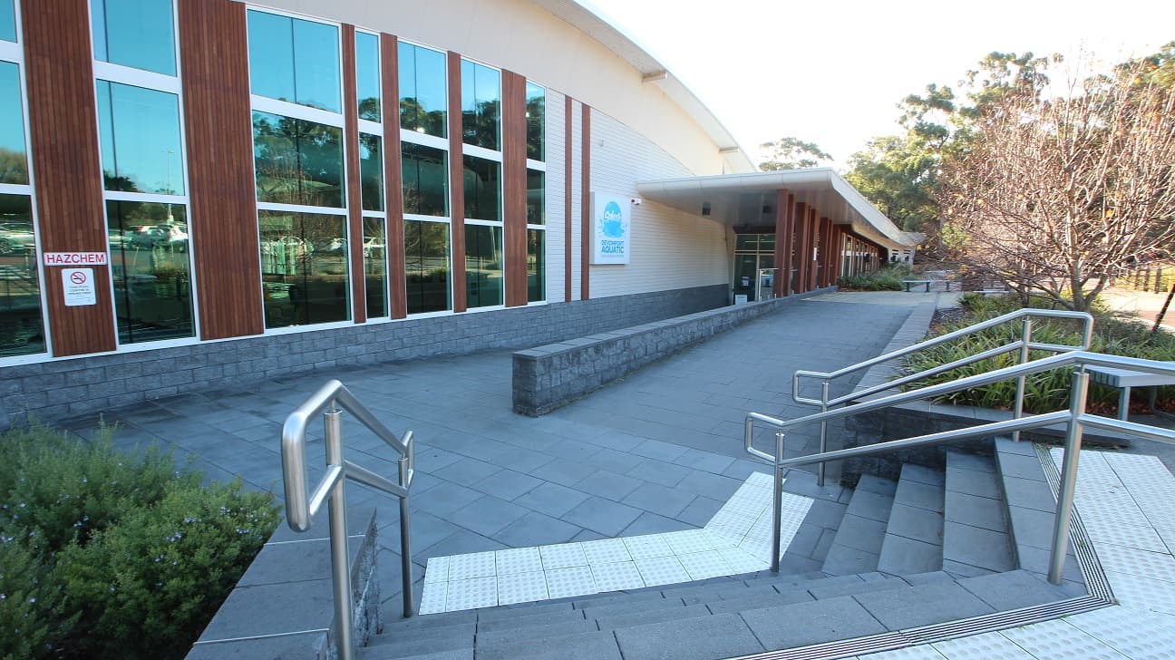 Staircase with metal handrails leading to one of the entrances of Splash Devonport. The floor is made of grey, square tiles. The walls of Splash Devonport are a combination of grey and white bricks, glass, and long stretches of wood arranged vertically. The glass walls reflect the car spaces and the surrounding area of the venue.