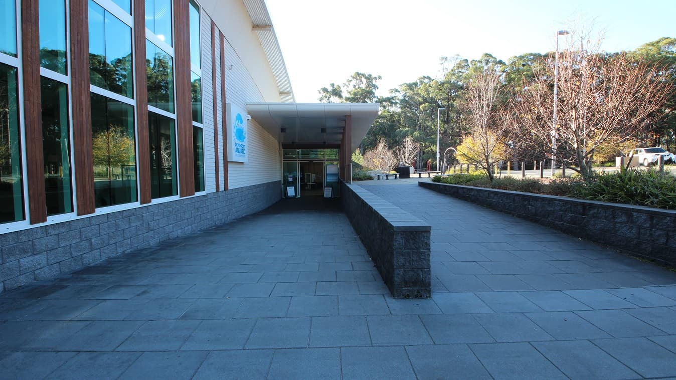 Accessible entrance ramp leading to the main entrance glass doors at Splash Devonport. Rectangular signboards are placed on the glass doors. On the left side wall is the colorful Splash Devonport Aquatic logo. Overlooking this section are tall trees with wide canopies that provide significant shade to the parking area.