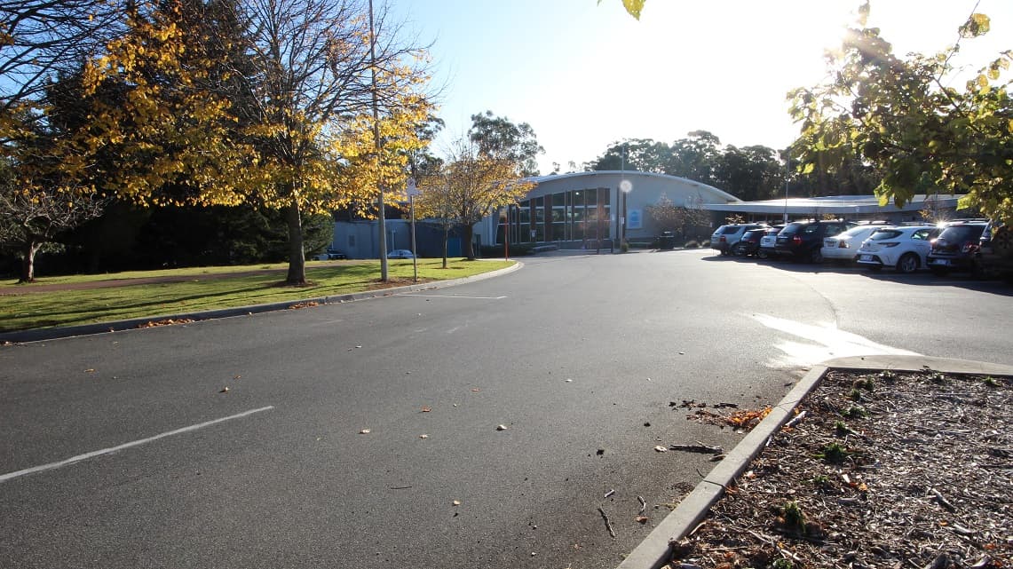 Road going to the Splash Devonport. On the right side is one of the accessible parking spaces near the entrance, while on the left side is a large space with Bermuda grass and trees with yellow leaves.