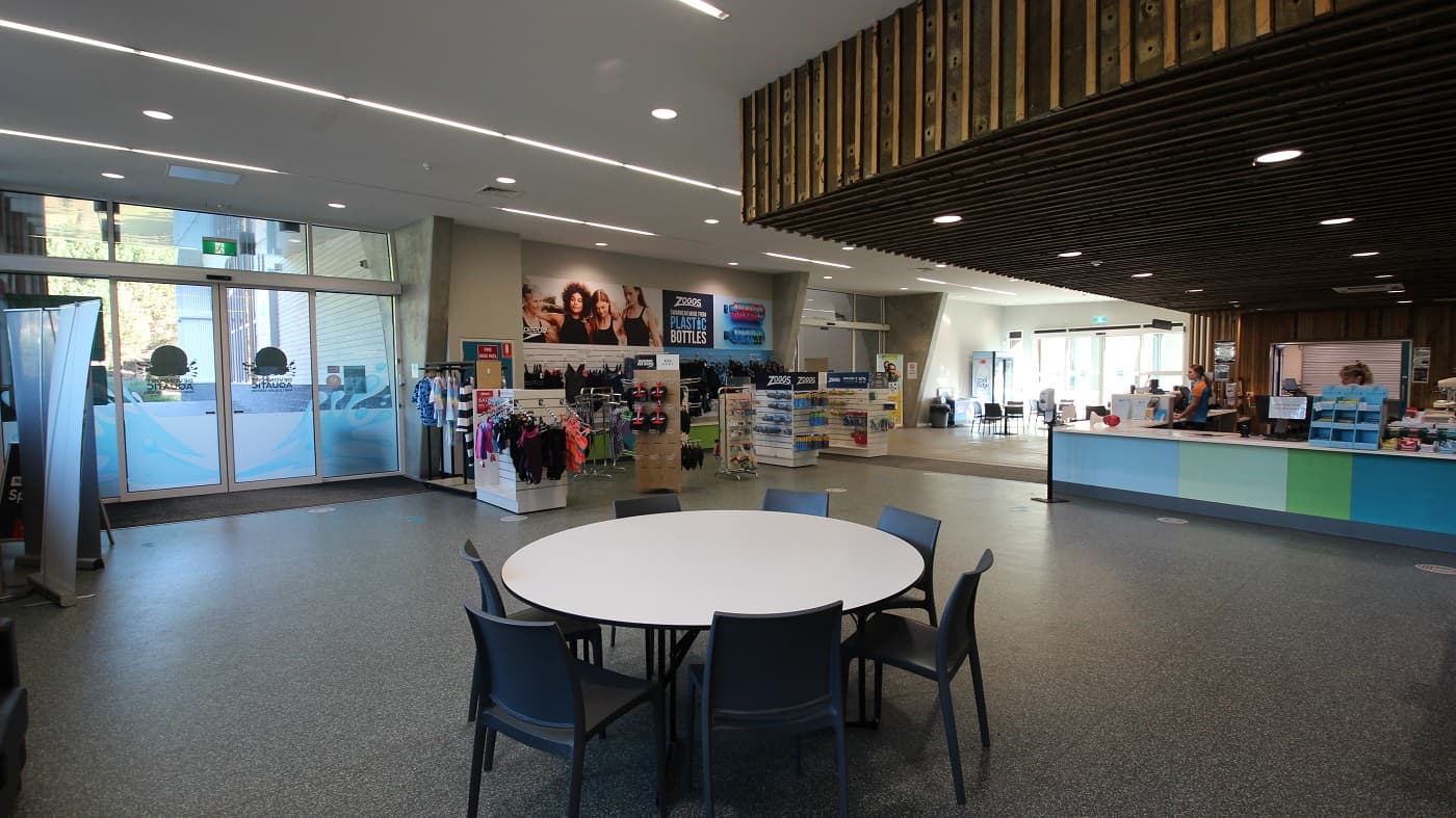 The seating area near the reception is composed of white circular tables with black plastic chairs. Aside from the swimming attire in the retail section are slippers and other swimming equipment. The reception desk is painted in different hues of blue and green color and is run by staff wearing orange and blue polo shirts.