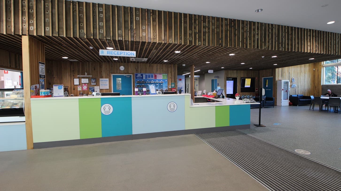 The reception area can be distinguished by the word reception hung on the wooden ceiling and a pattern of parallel lines in front of the reception area. The reception countertop is painted in different hues of green and blue. On the left side is the café, while on the right side is a liquid soap and alcohol dispenser in the reception area.