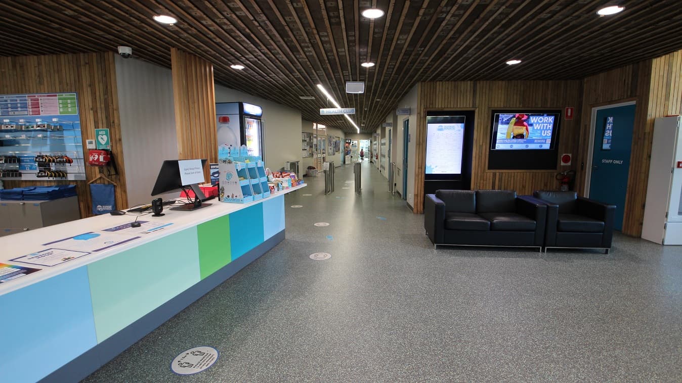 Heading straight from the main entrance doors and past the reception area is the corridor leading to the Health Club. The entry point to the Health Club is by the grey metal turnstiles. Opposite the reception desk are a staff-only blue-green door, black sofa chairs, and a television mounted on the wooden walls.