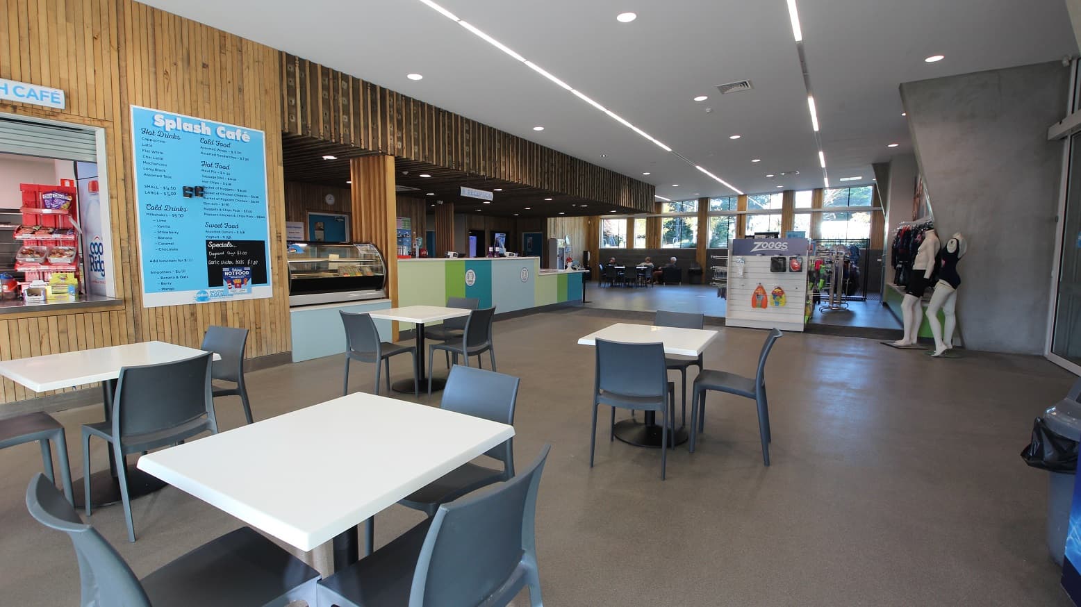Splash Café menu is typewritten on a poster fixed on the wooden wall beside the display stand on the sky blue café table countertop. The seating area of the café is composed of grey plastic chairs and white square tables.