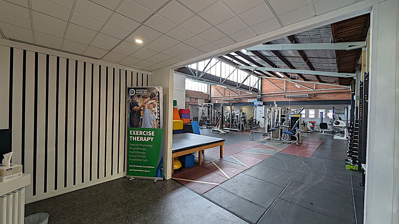 A gym interior with a focus on exercise therapy. On the left, there's a vertical striped wall with a tissue box on a small counter. A banner promoting exercise therapy stands next to a padded table. The right side features various gym equipment, including weight machines and exercise bikes. The floor is covered with rubber mats, and the ceiling has exposed beams and skylights, allowing natural light to illuminate the space.
