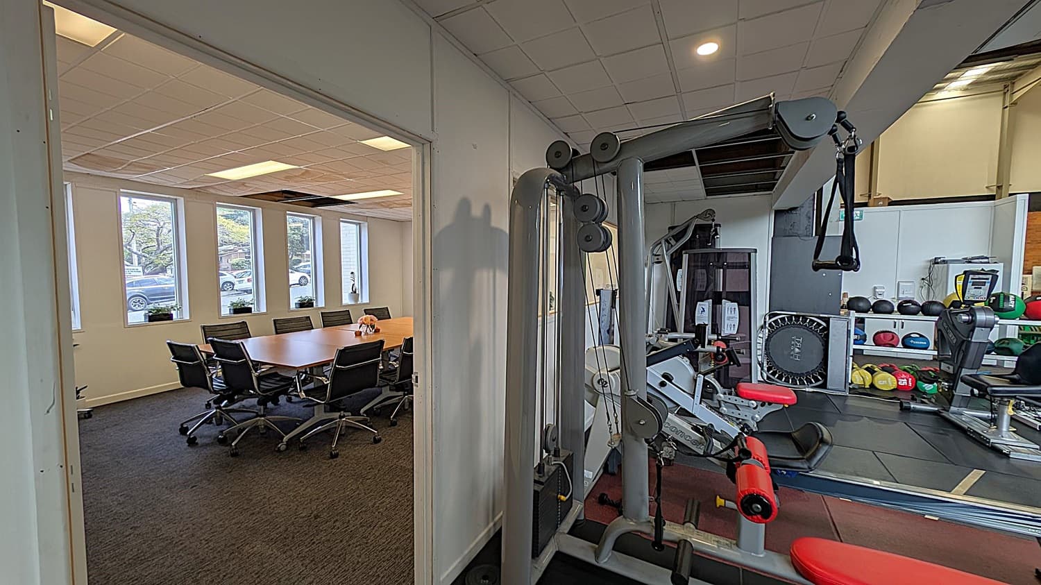 A room with a conference table and several black office chairs on the left, with large windows showing parked cars outside. On the right, a gym area with exercise equipment, including a weight machine, a small trampoline, and shelves with medicine balls. The floor is carpeted in the conference area and rubber in the gym. The ceiling has recessed lighting and a grid pattern.