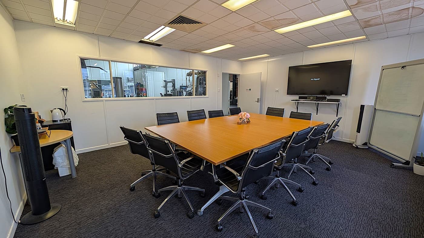 A conference room with a large rectangular wooden table surrounded by black office chairs on wheels. On the right wall, a large flat-screen TV displays "No Signal" above a shelf with a soundbar. A whiteboard stands nearby. On the left, a small round table holds a kettle and other items. A window in the back wall reveals an industrial area. The ceiling has fluorescent lights, and the floor is carpeted in dark grey.