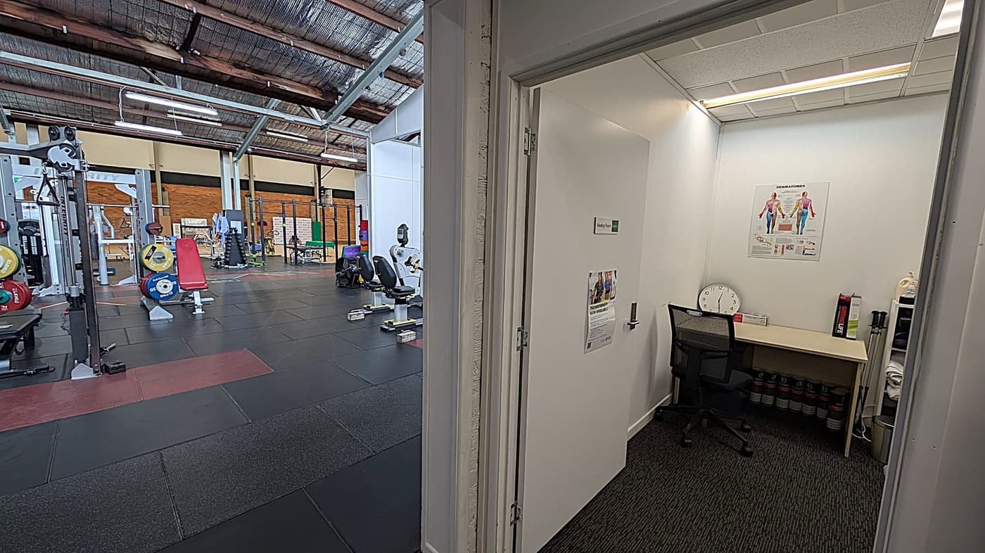 On the left, a gym with various equipment including weightlifting plates, a red bench, and exercise machines on a dark rubber floor. The ceiling is high with exposed beams and lighting. On the right, a small office with a desk, chair, and a wall poster showing human anatomy. A clock and some folders are on the desk, and the floor is carpeted. The ceiling has fluorescent lights.