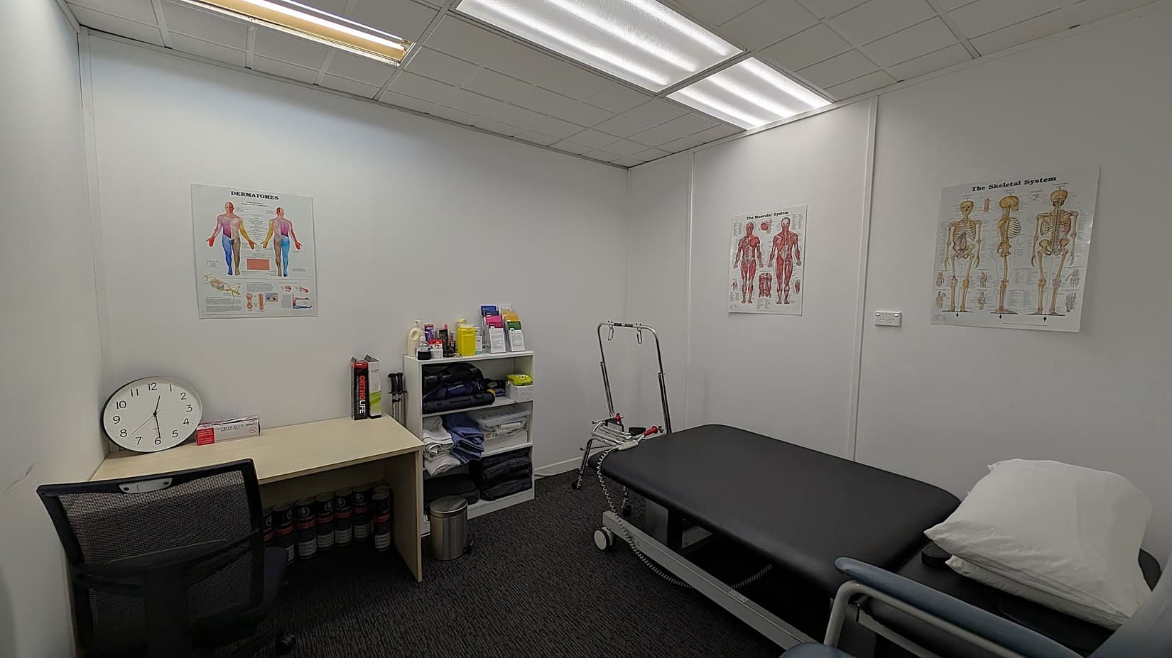 A small medical examination room with white walls and a dark carpeted floor. On the left, there's a desk with a large clock, a chair, and some medical supplies. In the centre, a shelving unit holds various bottles and pamphlets. On the right, a black examination table with a white pillow is positioned. The walls display anatomical posters. The ceiling has fluorescent lighting.