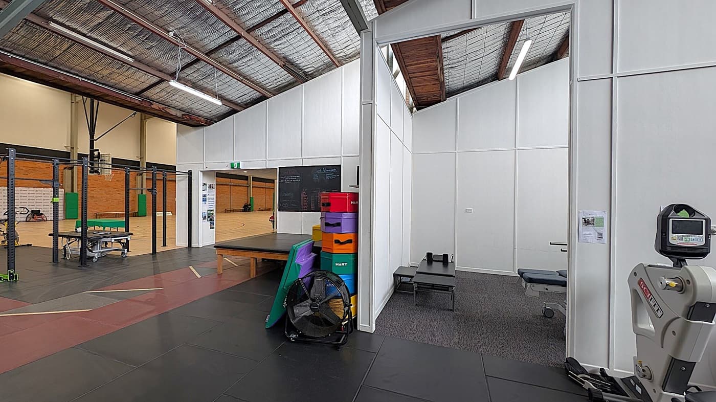 A gym interior with a high, exposed ceiling. On the left, there's a basketball court with a hoop and a brick wall. In the centre, a white partition wall separates the court from a workout area. Colourful exercise boxes and a large fan are in front of the partition. On the right, a small room with a grey carpet and a bench is visible. A piece of gym equipment with a digital display is in the foreground. The floor is covered with black and red mats.