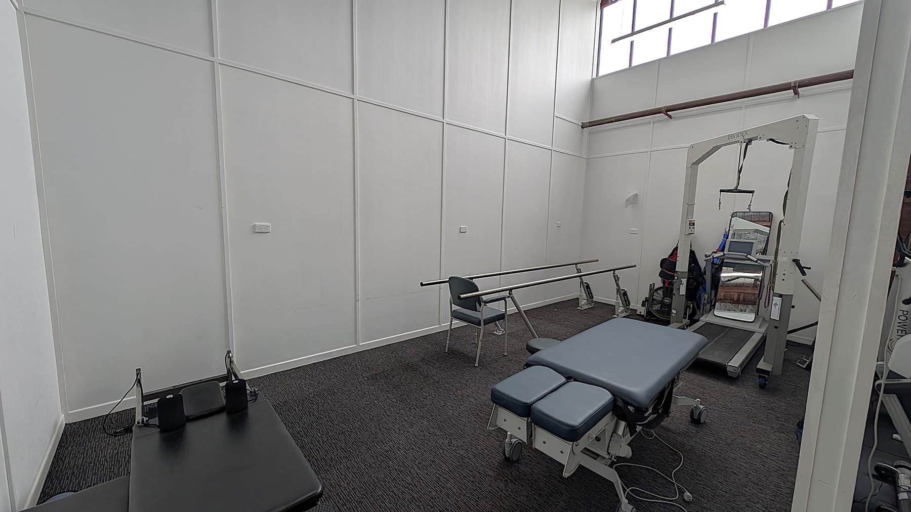 A room with white panelled walls and a high ceiling. On the right, there's a large exercise machine with a mirror. In the centre, a padded therapy table with wheels is visible. To the left, a chair is positioned next to parallel bars. The floor is covered with dark carpet, and there are high windows near the ceiling allowing natural light in.