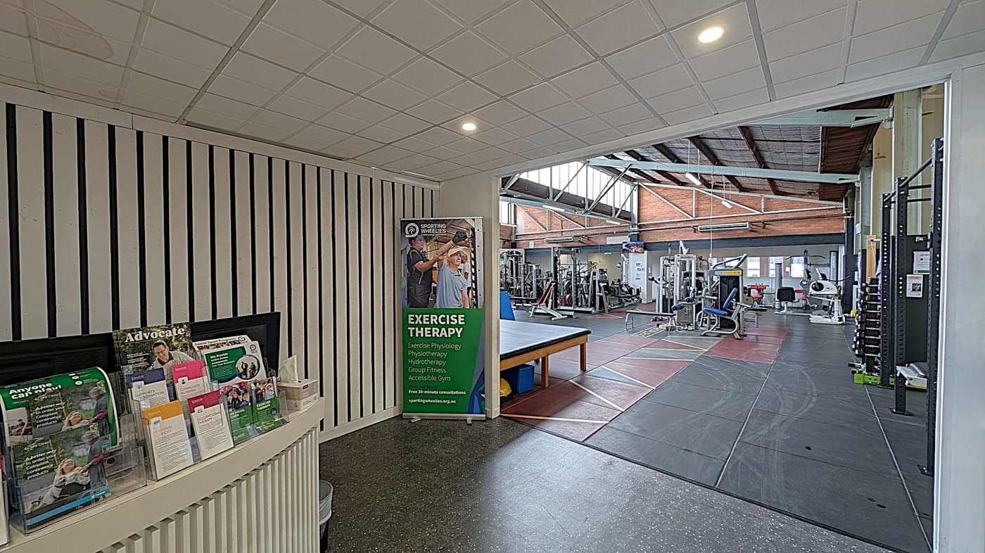 On the left, a counter displays brochures and magazines, with a box of tissues nearby. A vertical banner promoting exercise therapy stands beside it. The right side reveals a gym area with various exercise machines and equipment. The floor is covered with rubber mats, and the ceiling features exposed beams and skylights, allowing natural light to illuminate the space.