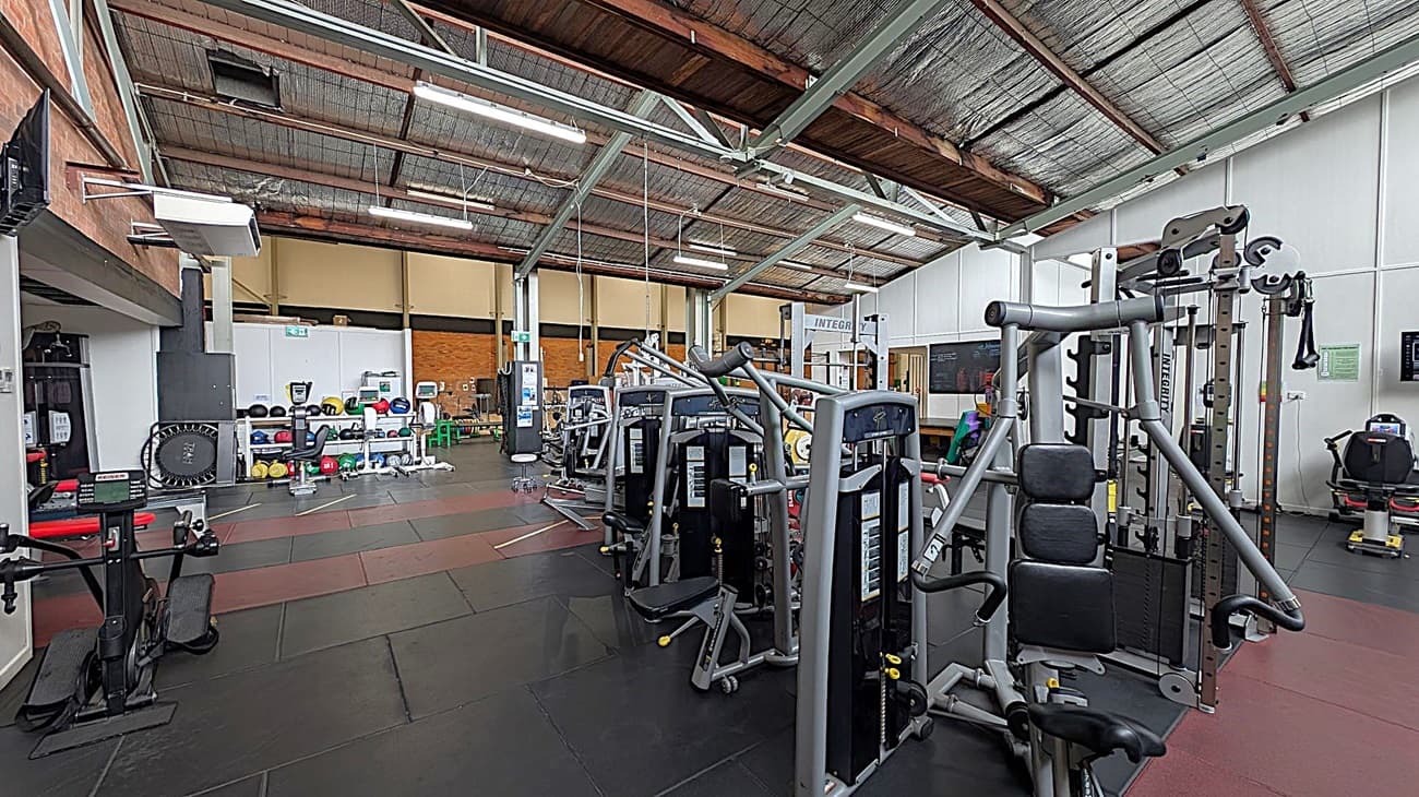 A gym interior with various exercise machines. On the right, there are weight machines and a leg press. In the middle, more weight machines are aligned. On the left, a trampoline and shelves with medicine balls and kettlebells are visible. The floor is covered with black and red mats, and the ceiling has exposed beams and lighting fixtures. The walls are a mix of brick and white panels.
