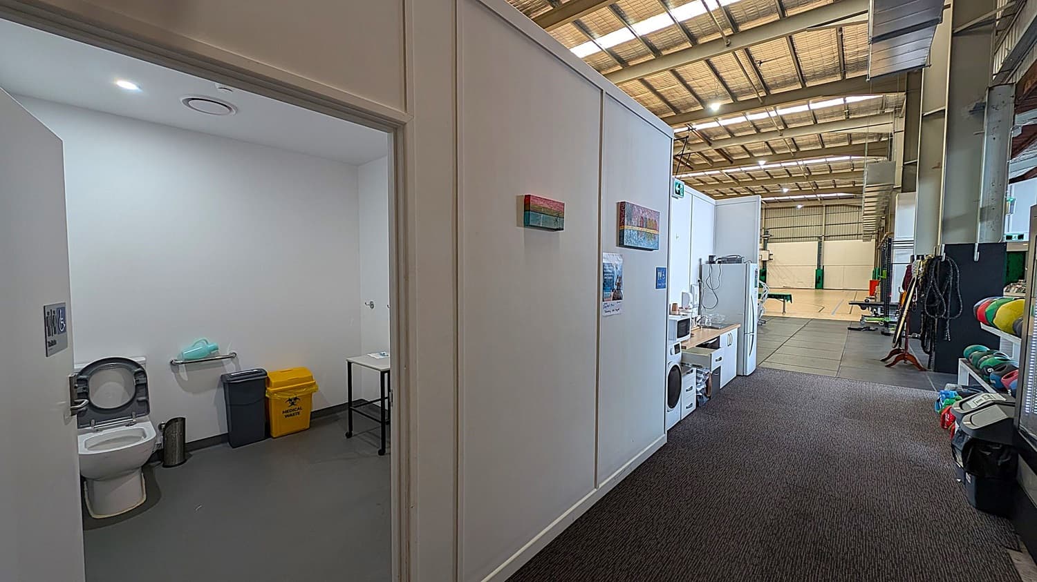 On the left, there's a bathroom with a toilet, a small table, and bins for general and medical waste. The middle section shows a corridor with white walls adorned with small paintings and a poster. On the right, there's a kitchenette area with a microwave and a washing machine. Further right, gym equipment and colourful mats are visible. The floor is carpeted, and the ceiling has exposed beams and lights.